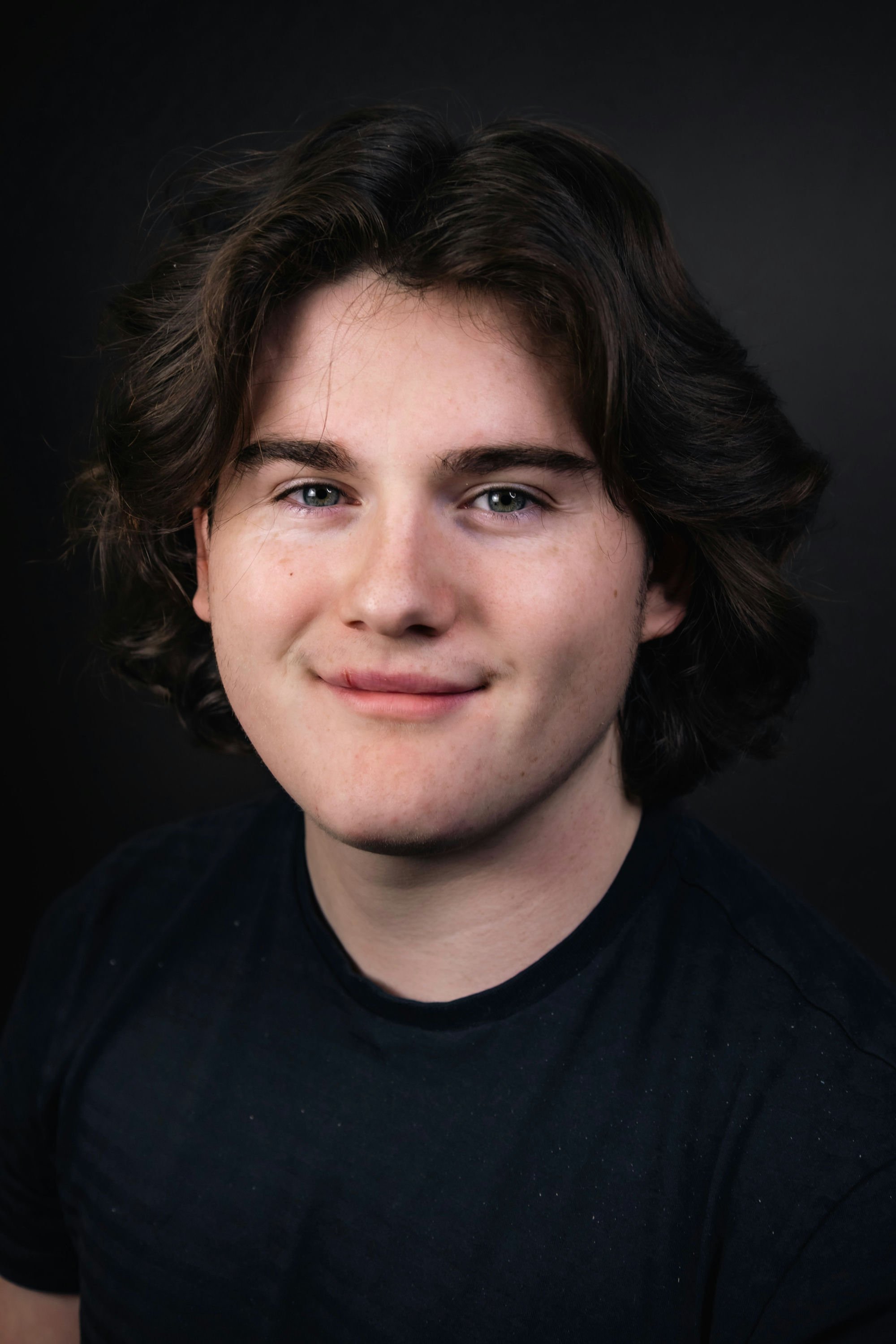 A woman with short, wavy blonde hair and blue eyes, wearing pearl earrings, a pearl necklace, and a sleeveless navy blue dress with thin white stripes, against a dark background.