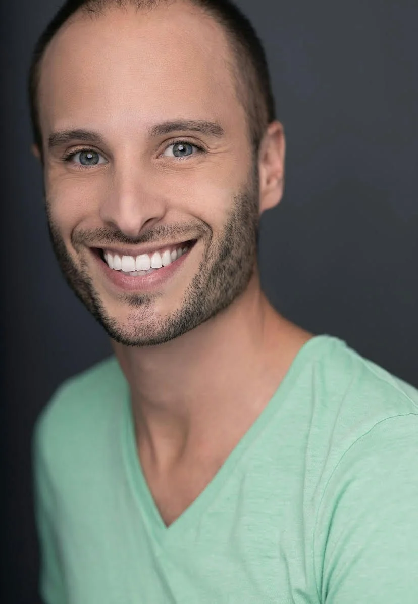 A woman with braided hair, wearing a blue sleeveless top, jewelry including earrings and necklaces, in an outdoor setting with blurred trees and grass in the background.