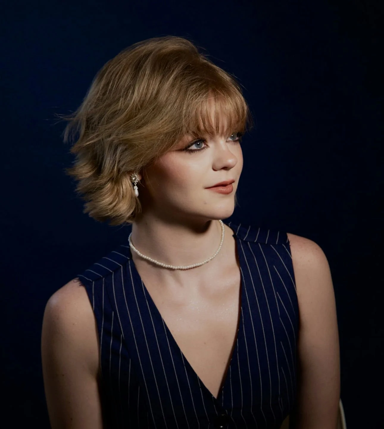 A woman with short, wavy blonde hair and blue eyes, wearing pearl earrings, a pearl necklace, and a sleeveless navy blue dress with thin white stripes, against a dark background.