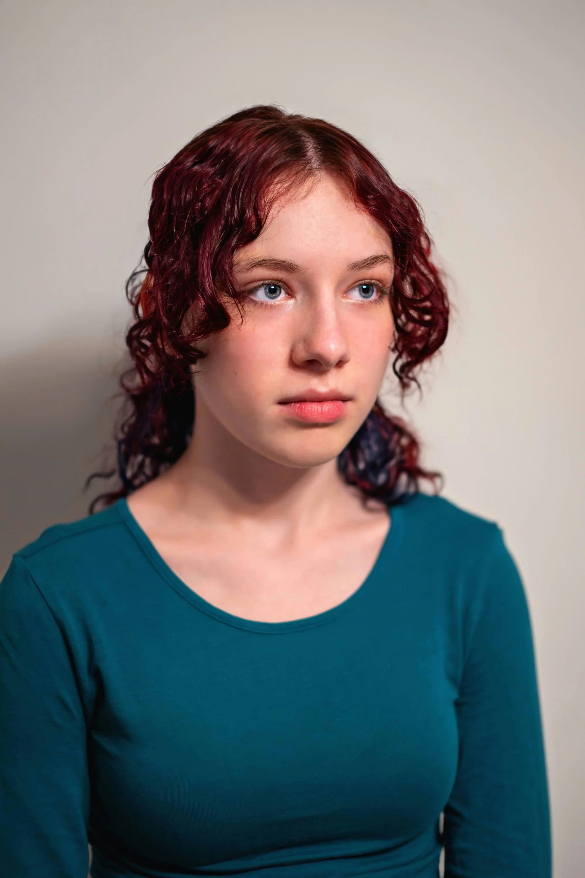 A young woman with curly red hair and blue eyes wearing a teal long-sleeve shirt, looking slightly to the side with a neutral expression, standing against a plain off-white background.