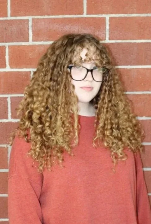 A young woman with long, wavy light brown hair and blue eyes, wearing a white sleeveless top, standing in front of a plain white wall, smiling softly at the camera.