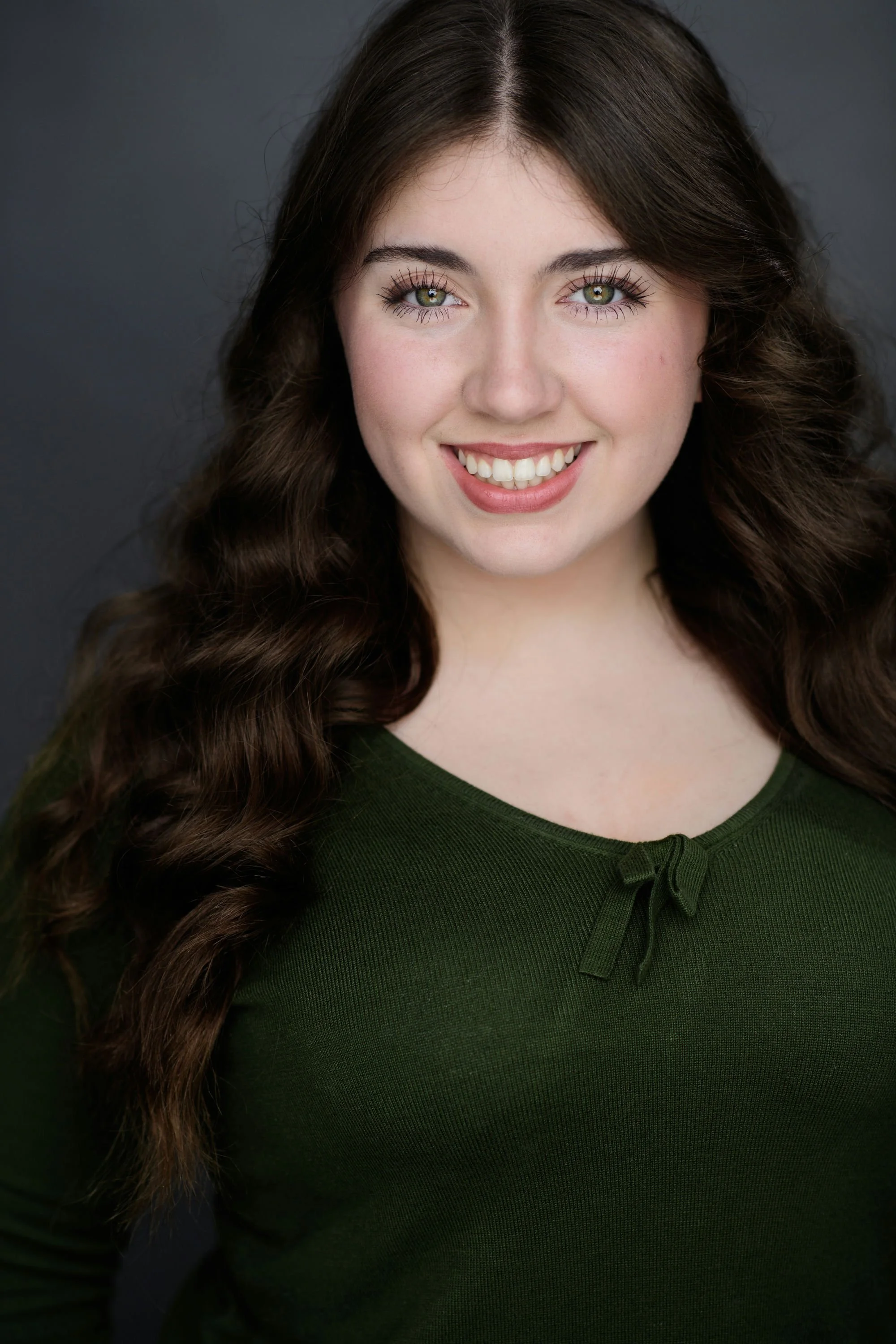 Close-up portrait of a young woman with long, wavy brown hair, green eyes, wearing a dark green top with a small bow at the neckline, smiling against a dark gray background.