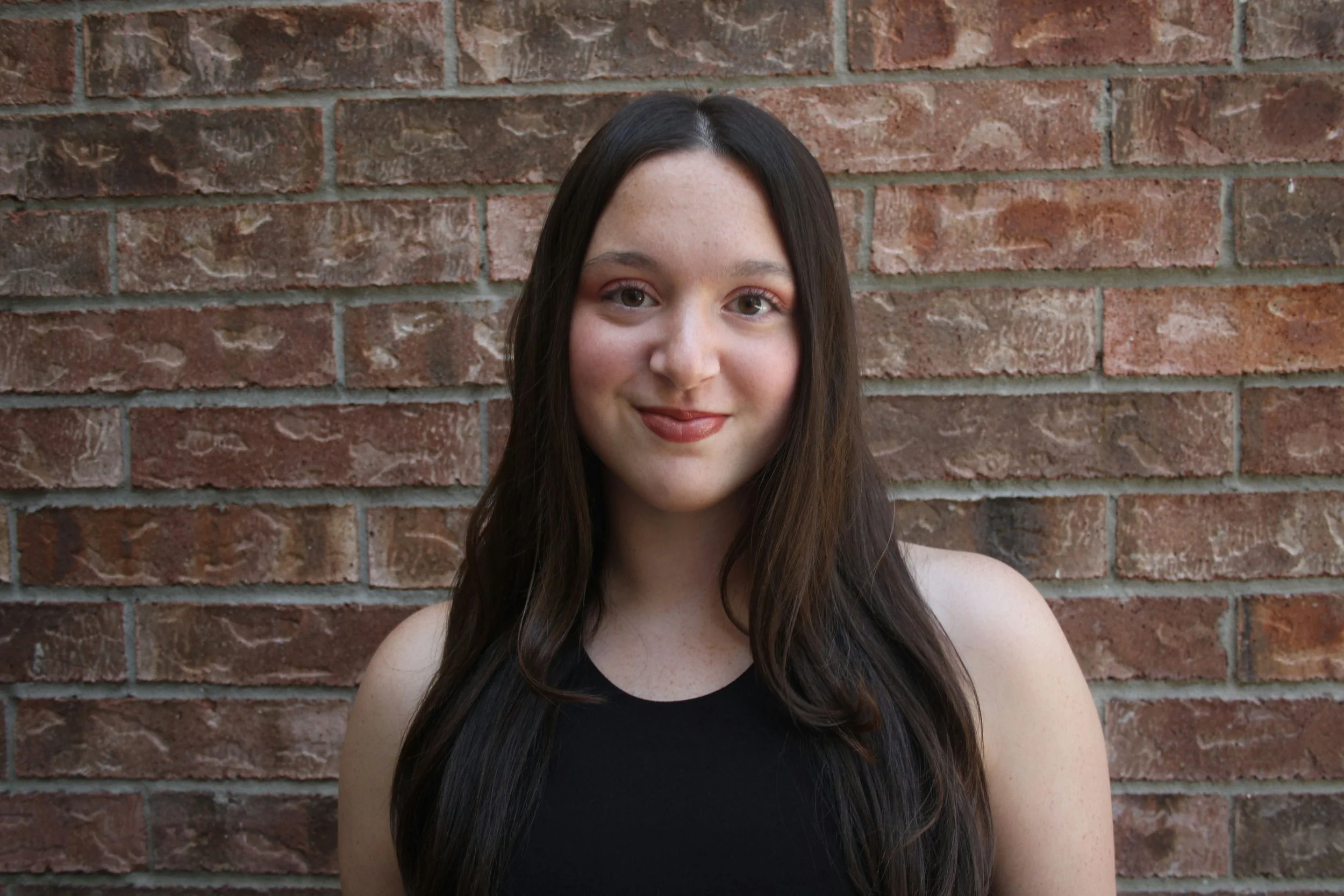 Young girl with long black hair, wearing a floral dress, standing outdoors in front of a wooden fence, smiling softly at the camera.