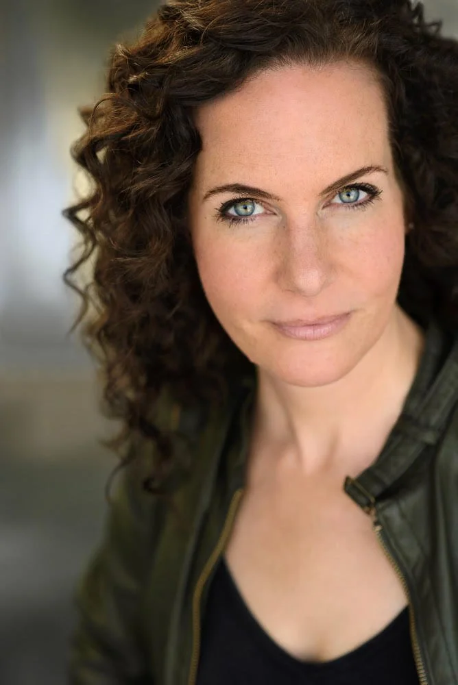 A woman with braided hair, wearing a blue sleeveless top, jewelry including earrings and necklaces, in an outdoor setting with blurred trees and grass in the background.