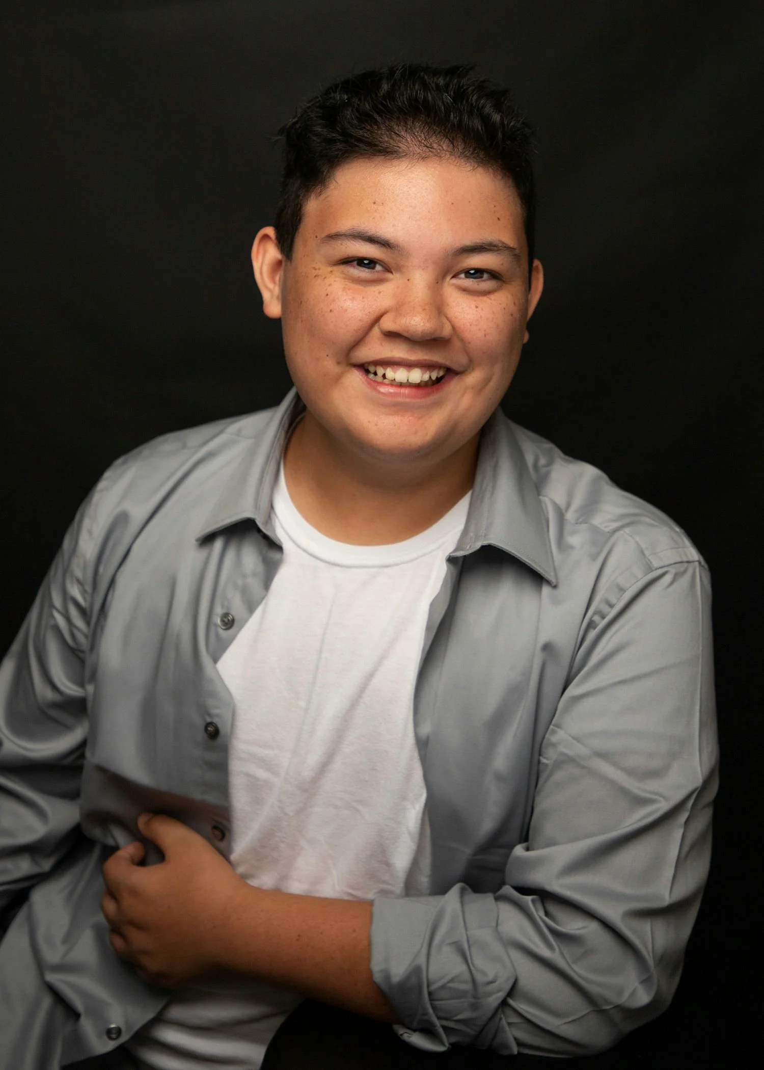 A smiling young person with short dark hair and freckles, wearing a light gray button-up shirt over a white t-shirt, posing against a black background.