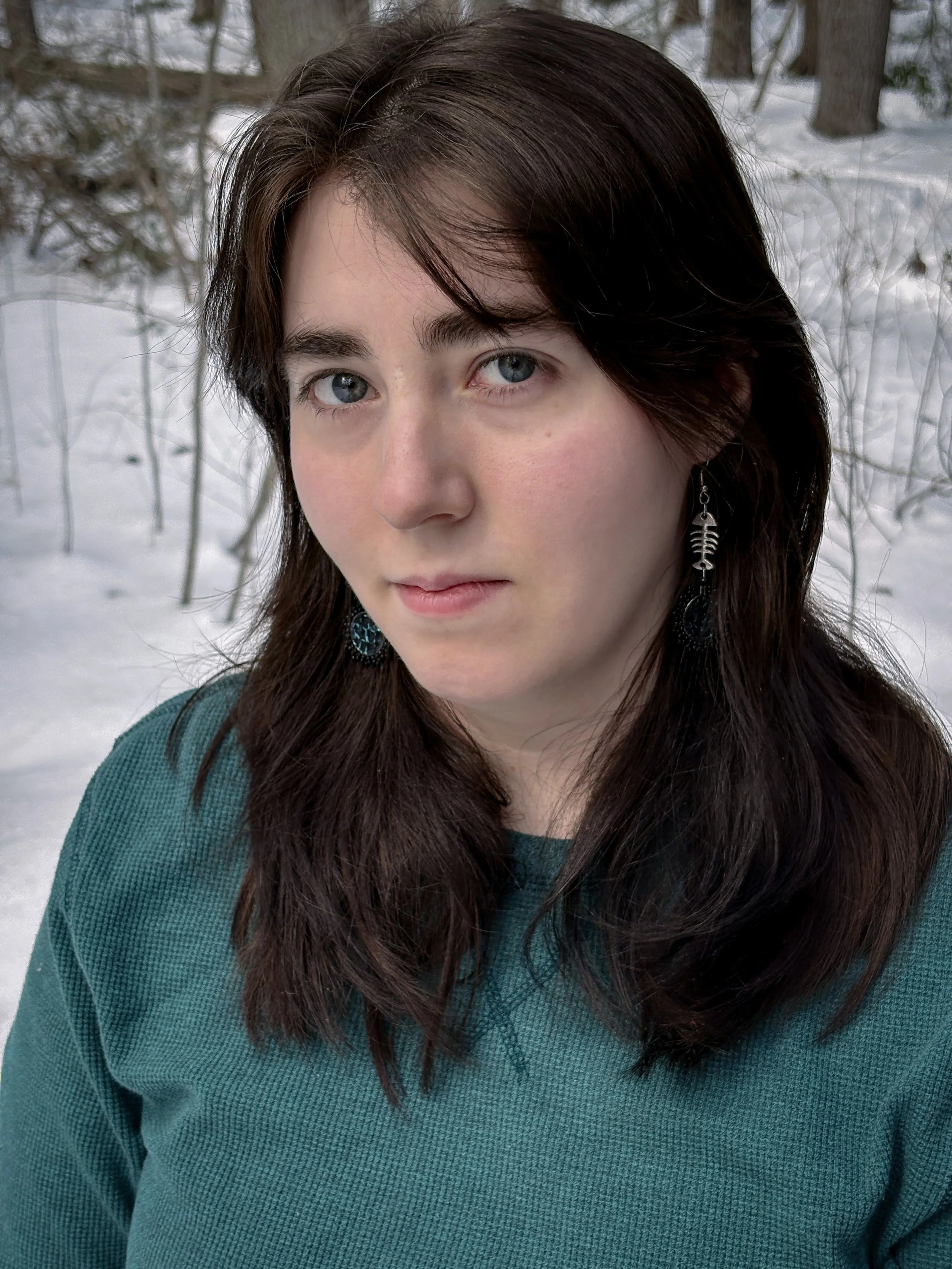A headshot of Mika McCrary with snow and trees behind her. She has long dark brown hair and wears a teal shirt and a fish bones earring.