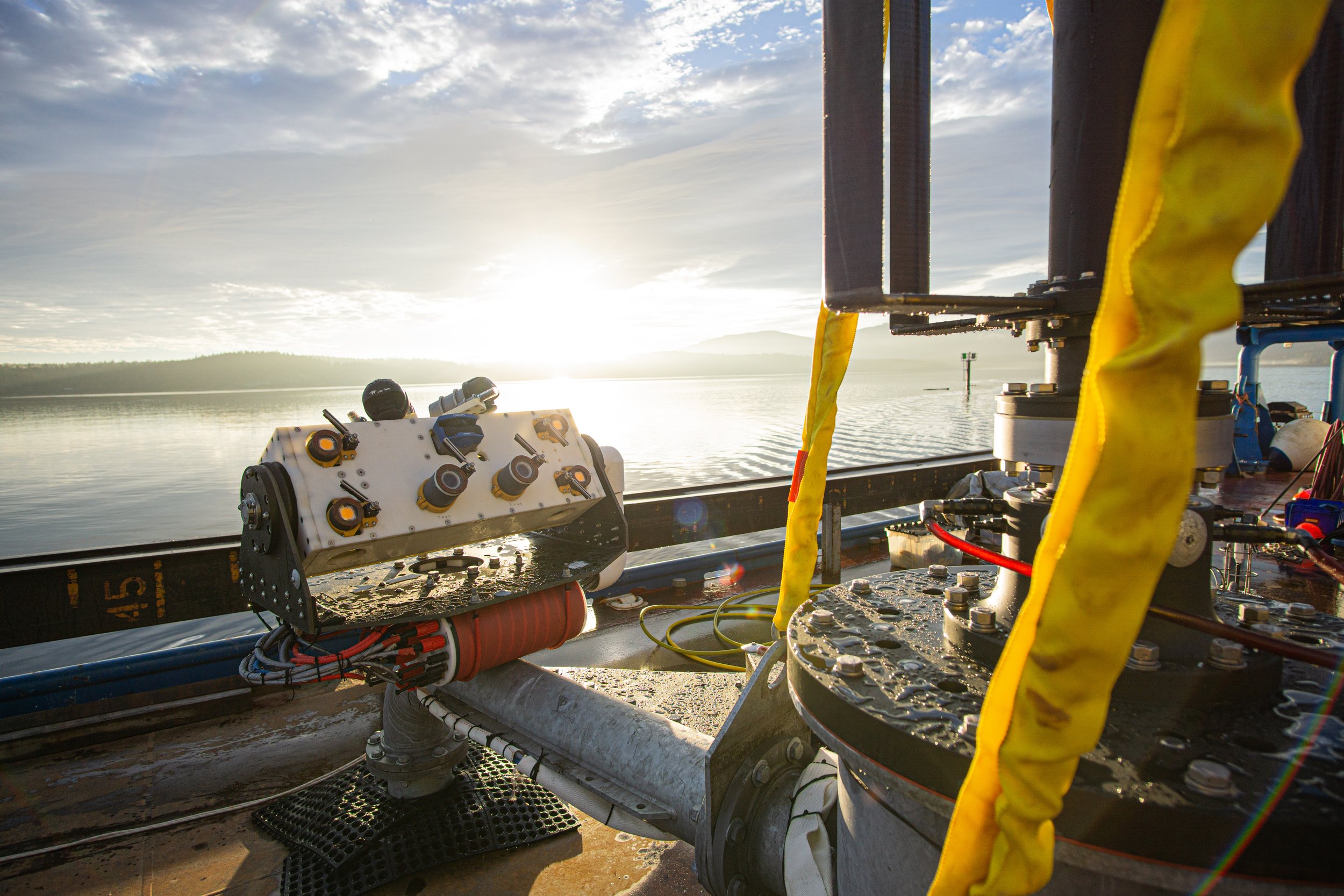The AMP, a rectangular box with multiple sensors, is mounted on the base of the Turbine Lander which is on the deck of a boat with the sun rising over Sequim Bay in the background.