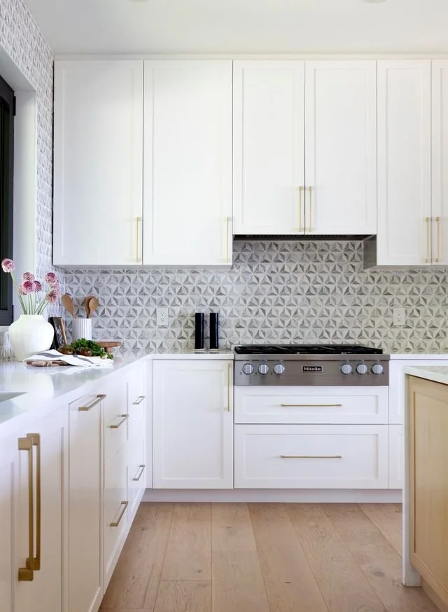 A modern white kitchen with upper and lower cabinets, gold handles, a patterned gray backsplash, a gray stove, a white countertop, a white vase with pink flowers, and some small kitchen tools.