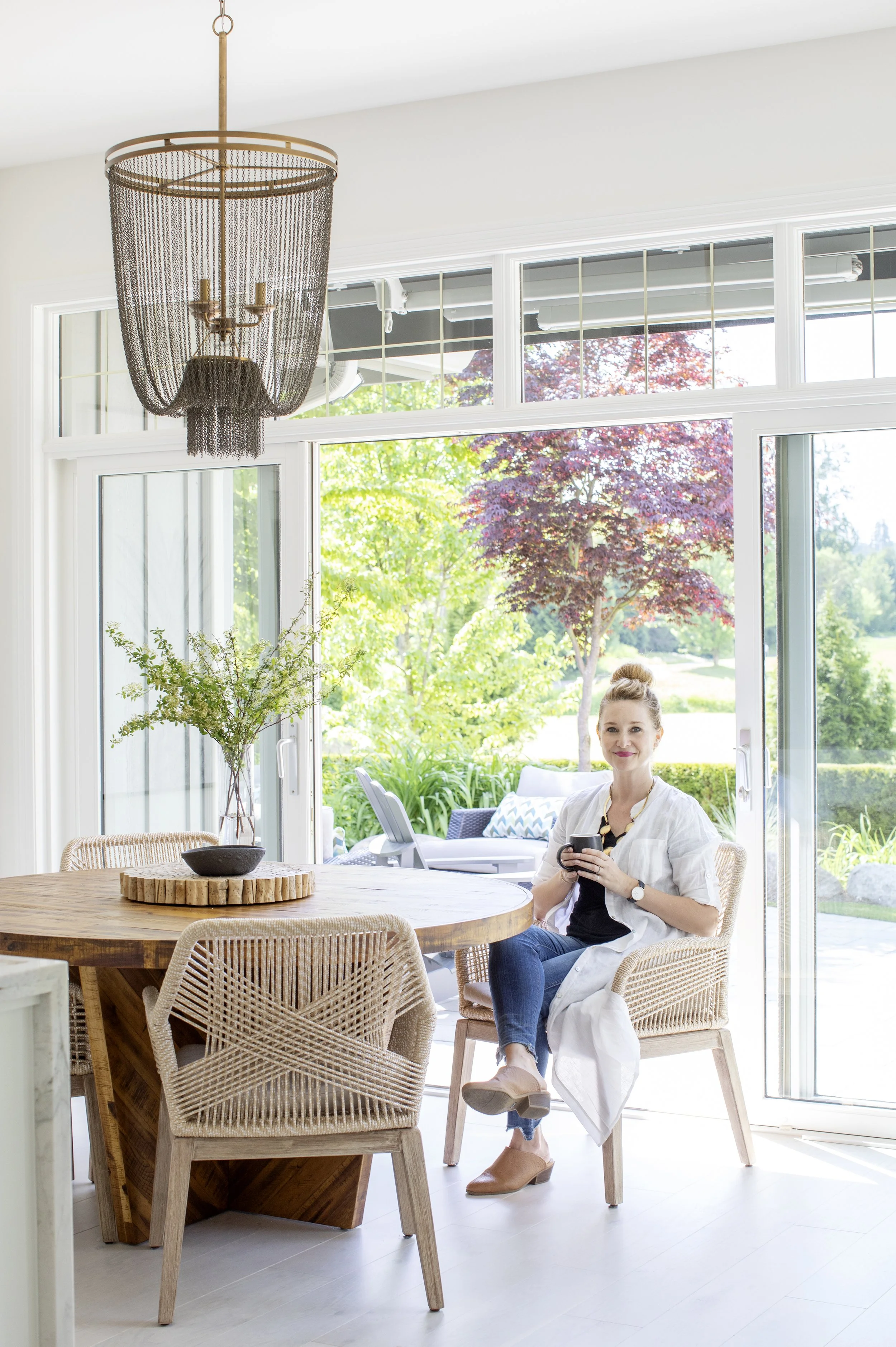 A woman sitting on a wicker chair at a round wooden dining table in a bright, open dining area with large sliding glass doors and greenery outside.
