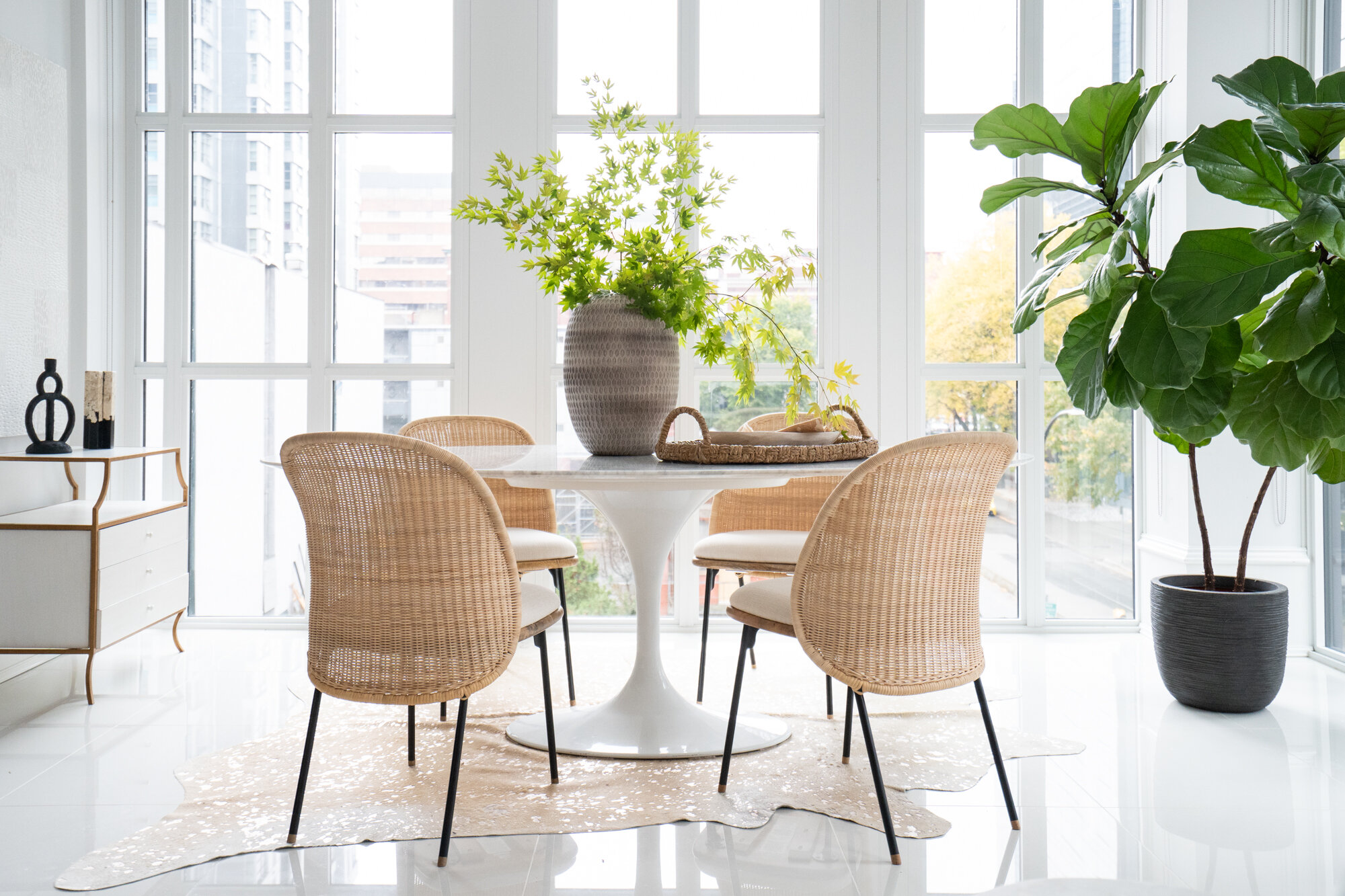 Bright dining area with a round white table, four wicker chairs with cream cushions, large potted plants, and a natural fiber rug beneath the table, with large windows in the background letting in natural light.