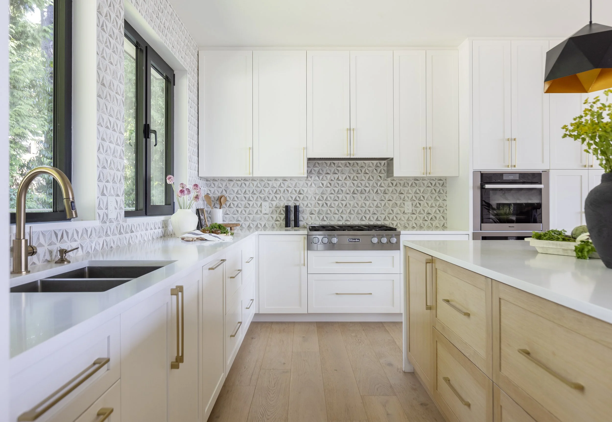 Modern bright kitchen with white and light wood cabinets, black window frames, and a patterned tile backsplash beneath white cabinets. There is a sink, decorative items, and a black and stainless stove. A kitchen island with a light wood finish is vi