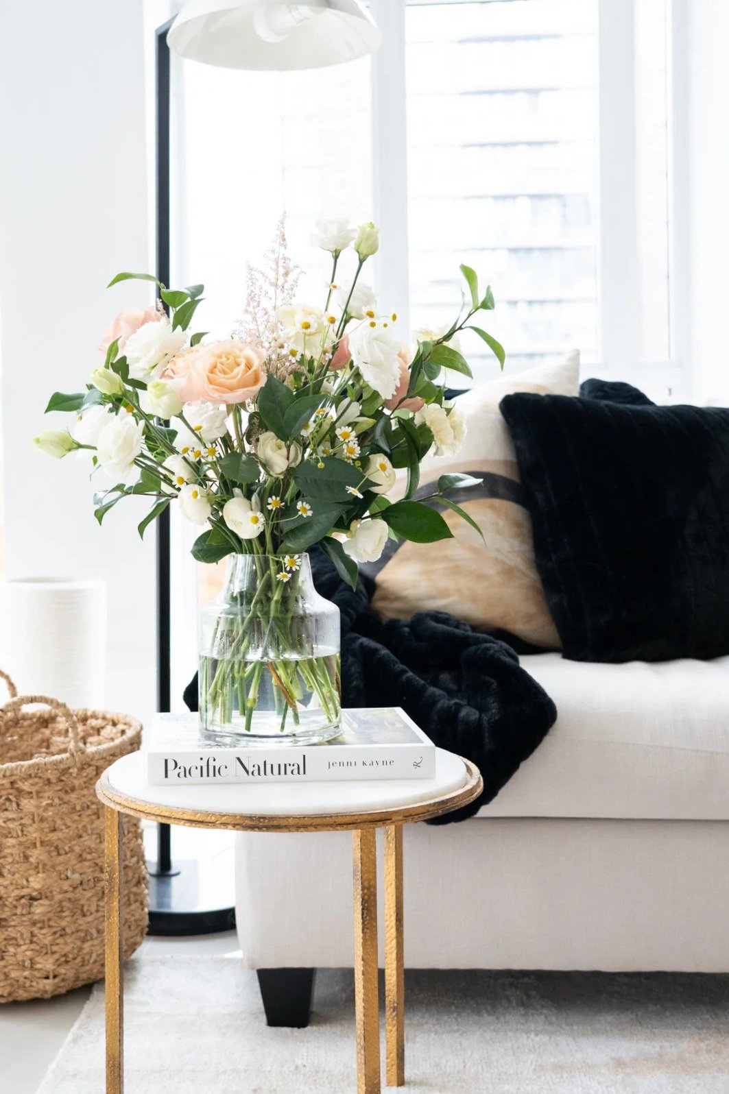 A glass vase with a bouquet of pink roses, white flowers, and greenery on a white book titled "Pacific Natural" on a gold side table inside a bright room with large windows.