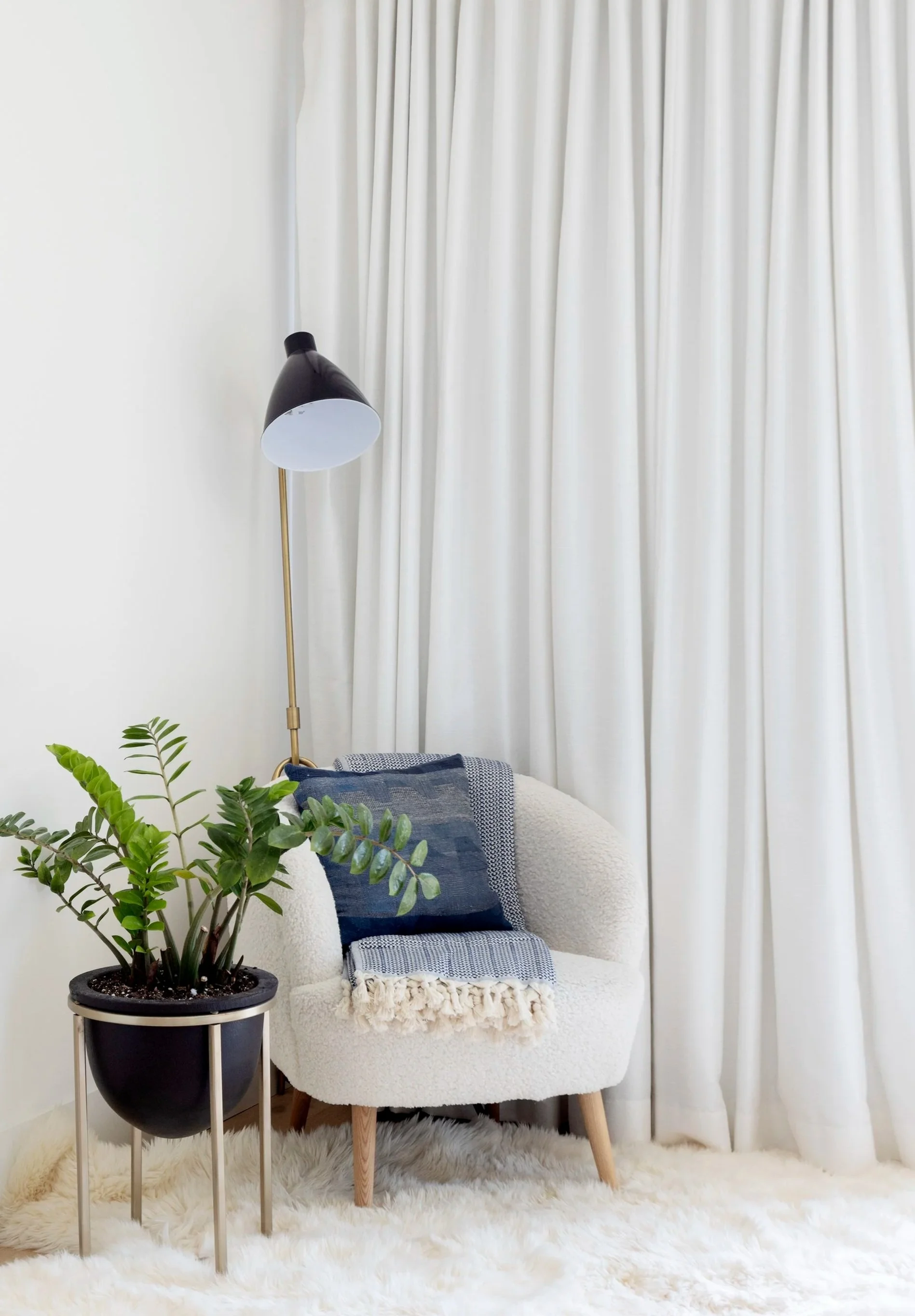 A cozy corner with a white textured armchair, a black potted plant on a stand, a blue pillow, a cream blanket, a tall black and gold floor lamp, white curtains, and a fluffy cream rug.