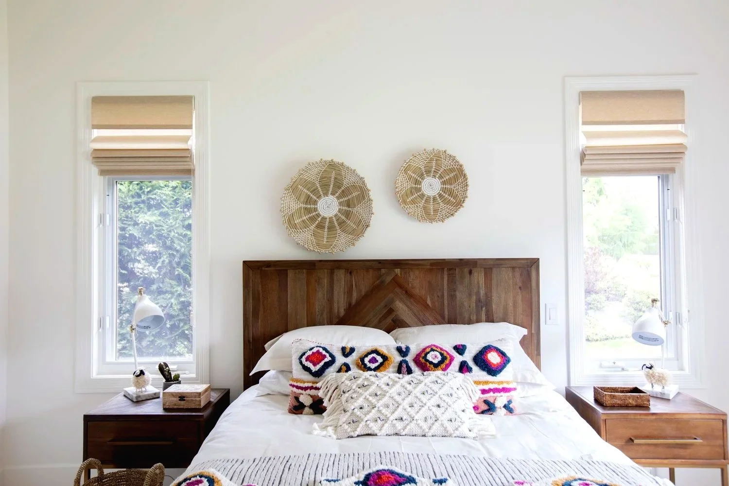 A bedroom with a wooden headboard, two nightstands, and two windows. There are decorative pillows on the bed with colorful knit patterns, and two woven wall hangings above the headboard.