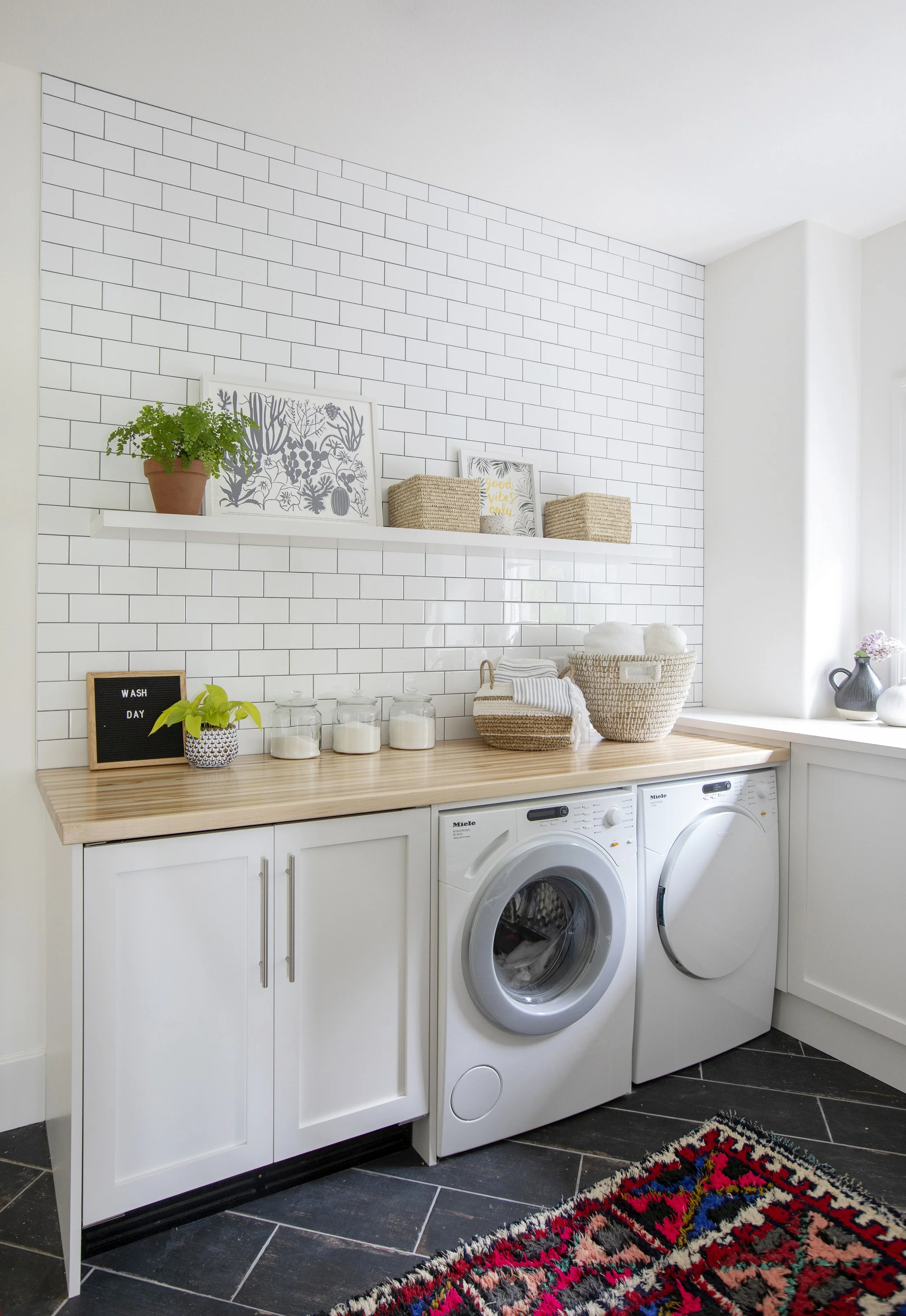 Laundry room with white cabinets, a wooden countertop, white tiled wall, a washing machine, a dryer, decorative plants, jars, baskets, and framed art.