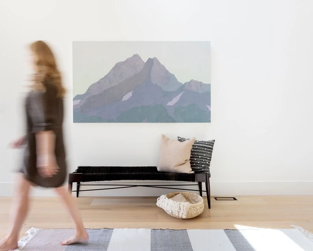 A blurred woman walking past a minimalist living room with a black bench, two pillows, a basket, a large mountain artwork, and a light-colored rug.