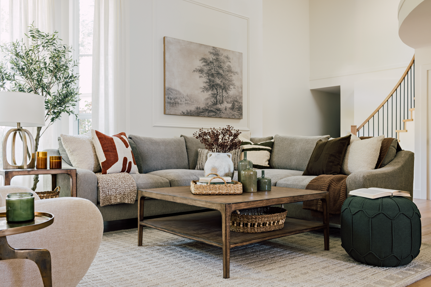 Living room with a gray sectional sofa, wooden coffee table, and various decorative pillows and vases, with a large window and staircase in the background.