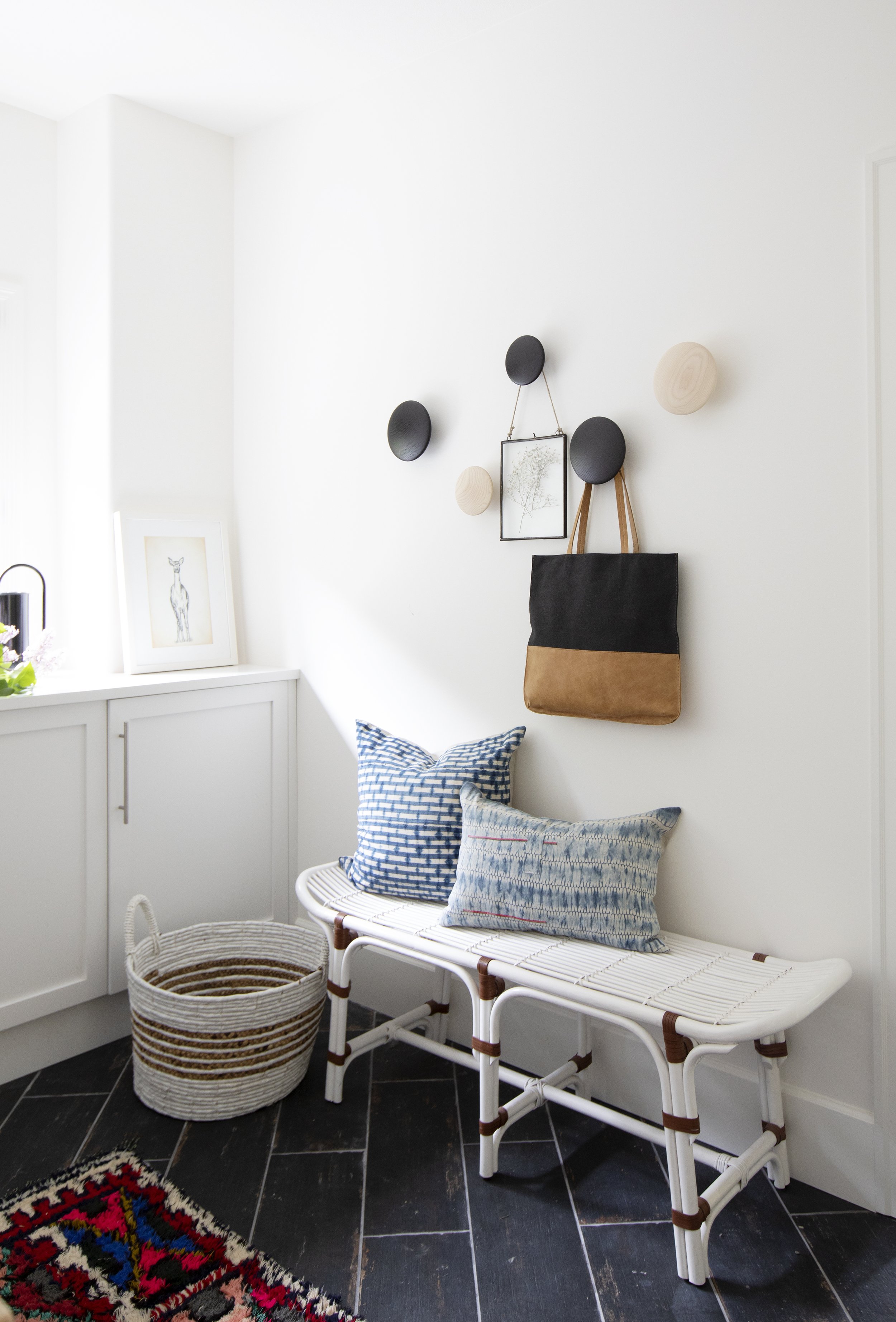 A white bench with two patterned pillows against a white wall decorated with circular wall hooks, a black and brown tote bag, and a small framed art piece. There is a white basket on the black floor next to a colorful rug.