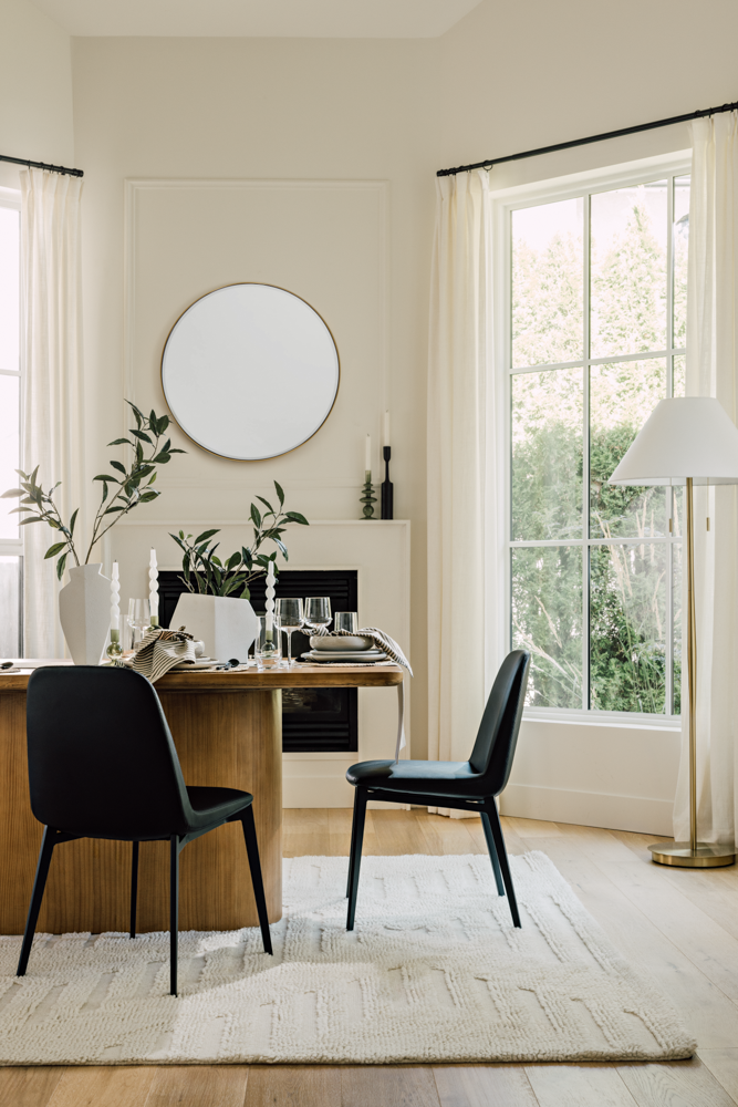A dining room with a wooden table, two black chairs, and white tableware, next to large windows with cream curtains, a white rug, a white floor lamp, and a white vase with green plants.