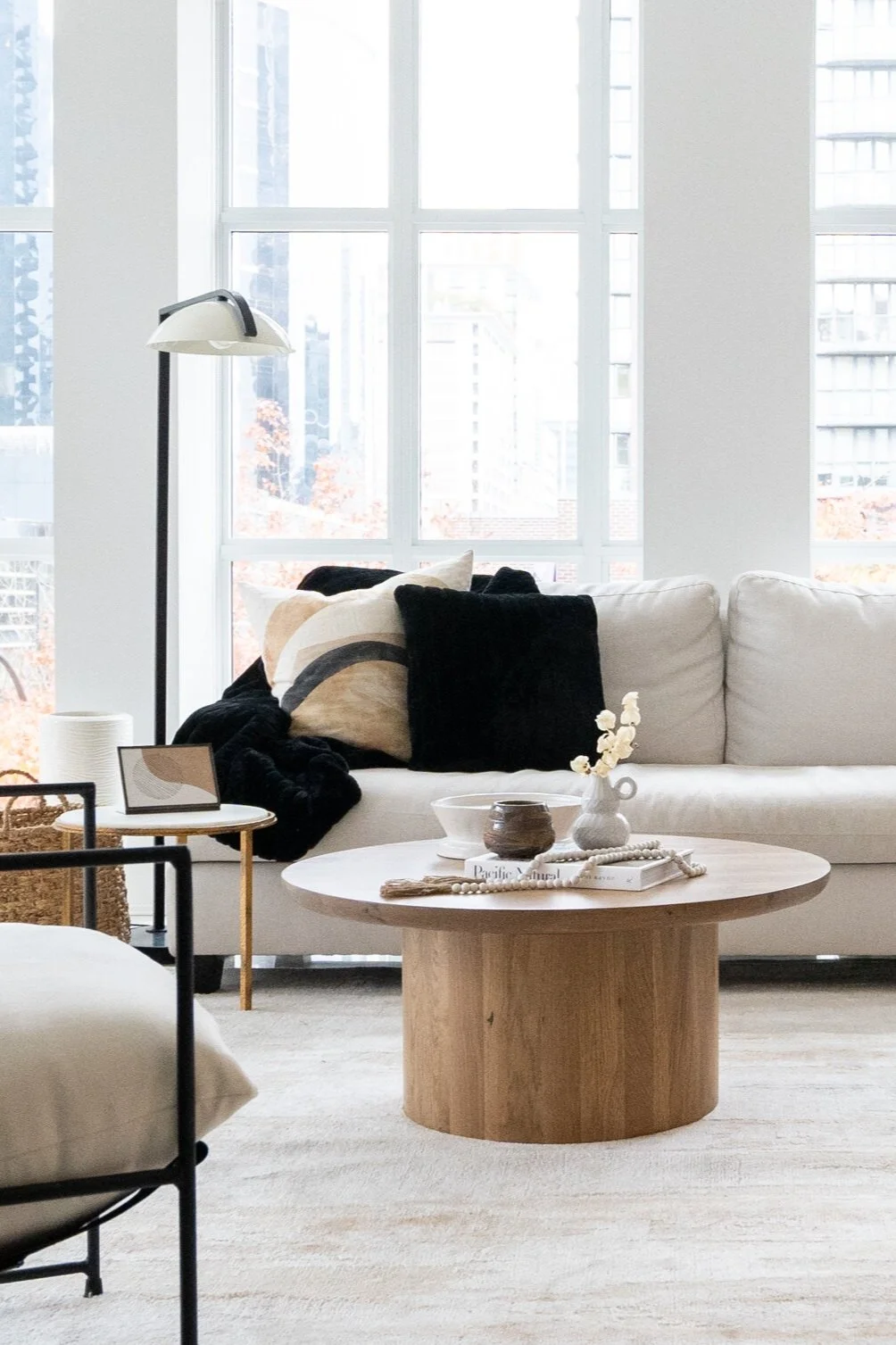 Living room with a white sofa, black and beige pillows, a small side table with decor, a round wooden coffee table with a vase and books, large windows letting in natural light, and a white floor lamp.