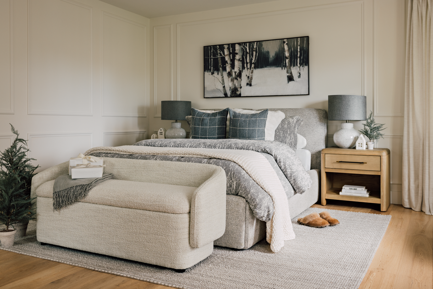 A cozy bedroom with a large bed, gray and white bedding, and dark blue pillows. There's a wooden nightstand with a gray lamp, small house decorations, and a small plant. A painting of a winter forest hangs above the bed. There is a beige textured ben