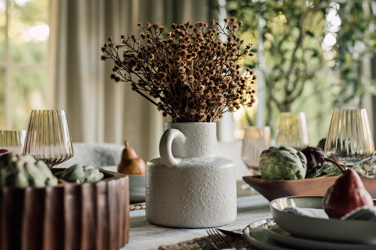 A table decorated with a textured white vase with dried flowers, placed among plates with artichokes and pears, and surrounded by glassware, with a window and greenery in the background.