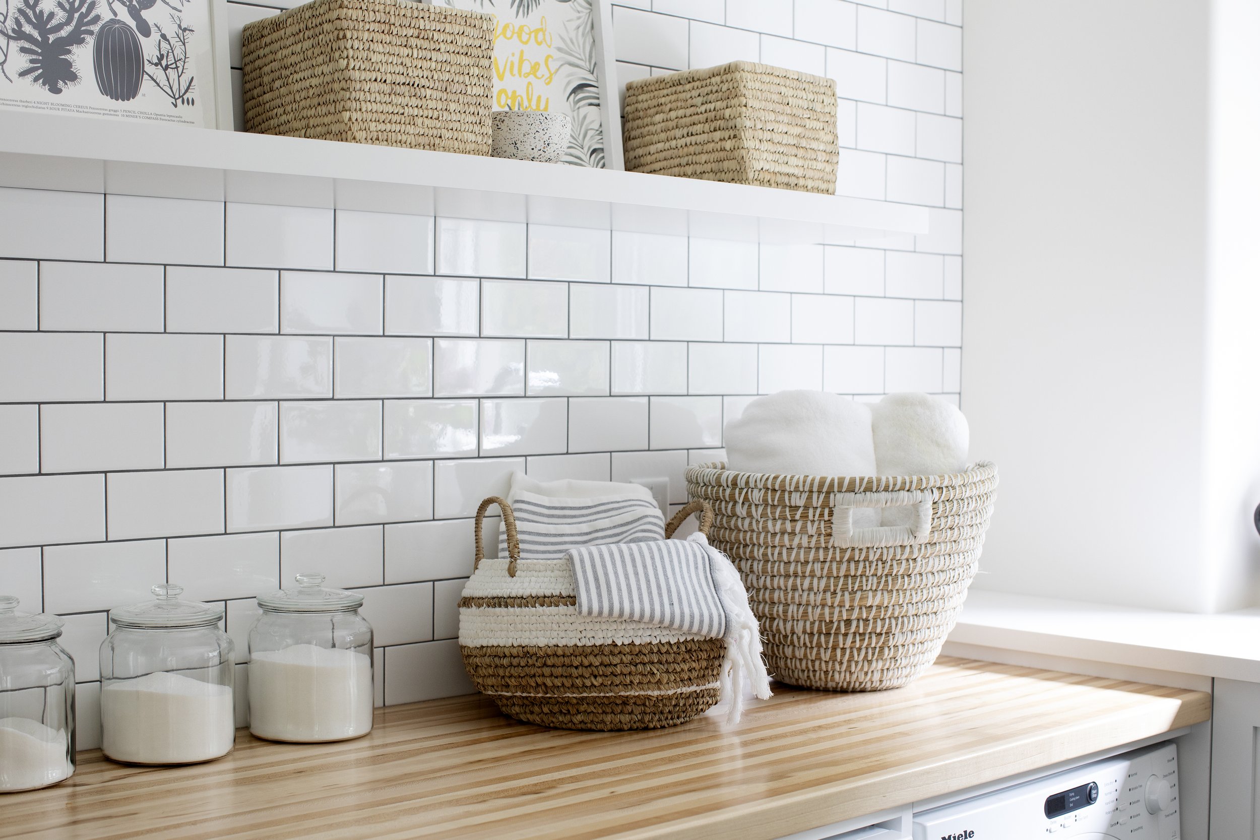 A wooden countertop with three glass jars filled with white powder, a striped towel resting on a woven basket, and a woven laundry basket filled with white rolled towels. White subway tile wall in background with a white shelf above holding various w