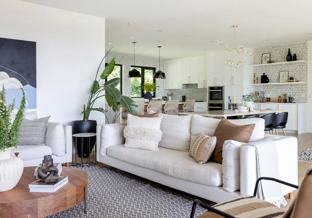 Open-concept living room and kitchen area with white furniture, large potted plants, and modern decor.