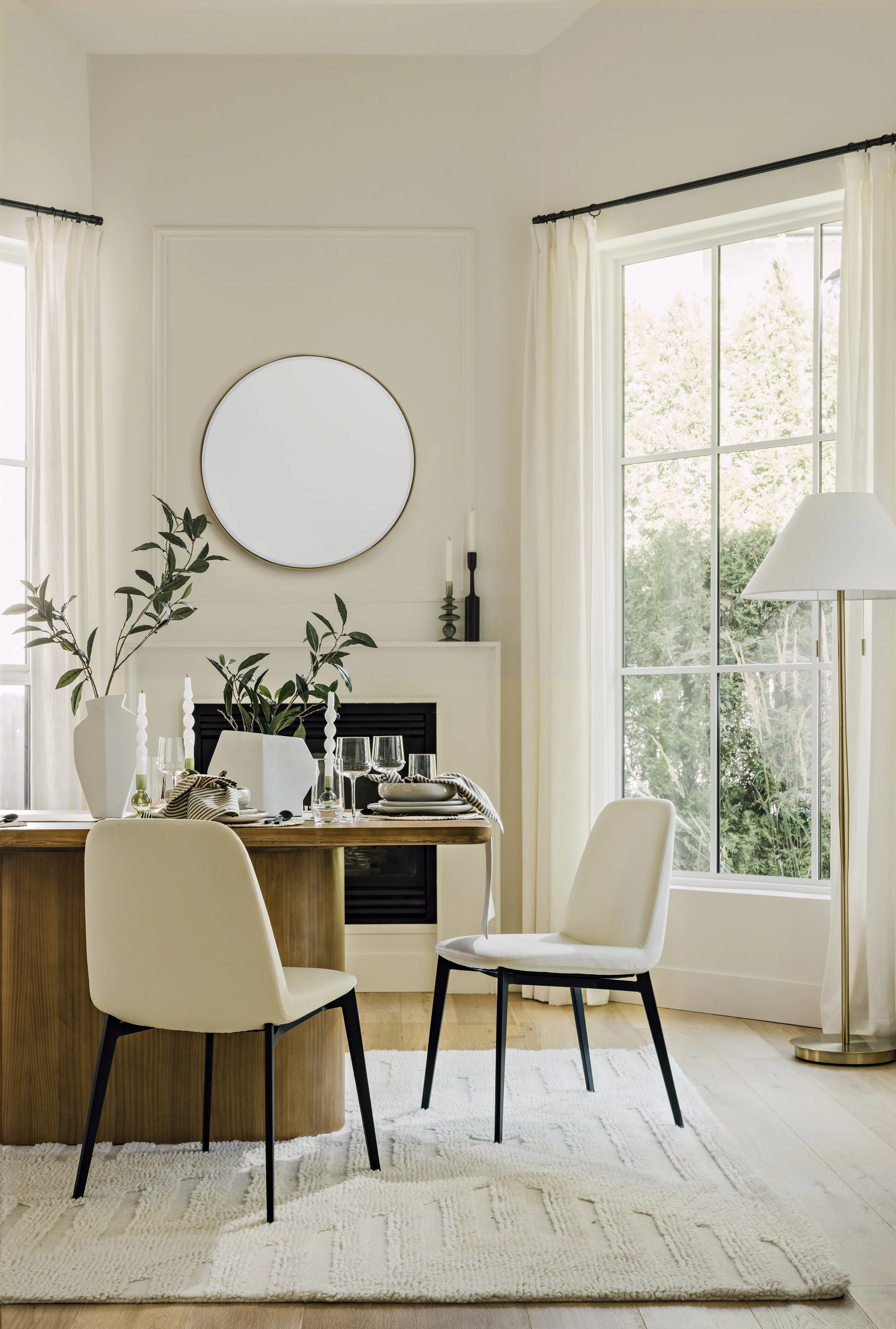 A bright dining area with a hardwood table, two white chairs, large windows with white curtains, and minimalistic decor including a round mirror, candles, greenery, and table settings.