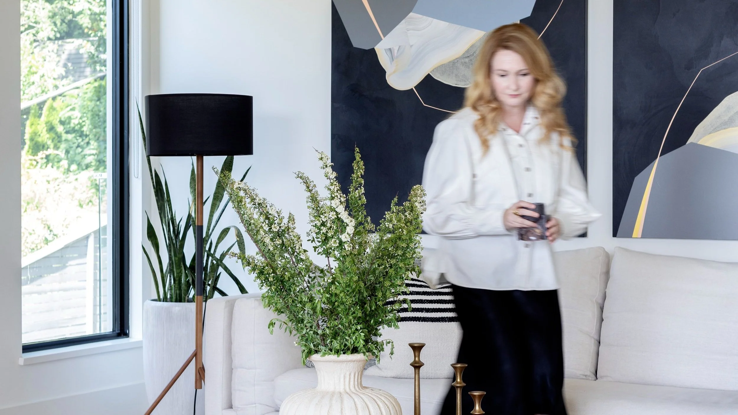 A woman with long red hair wearing a white blouse and black pants holding a smartphone, standing in a modern living room with a white couch, large potted plant, black floor lamp, and abstract art on the wall.