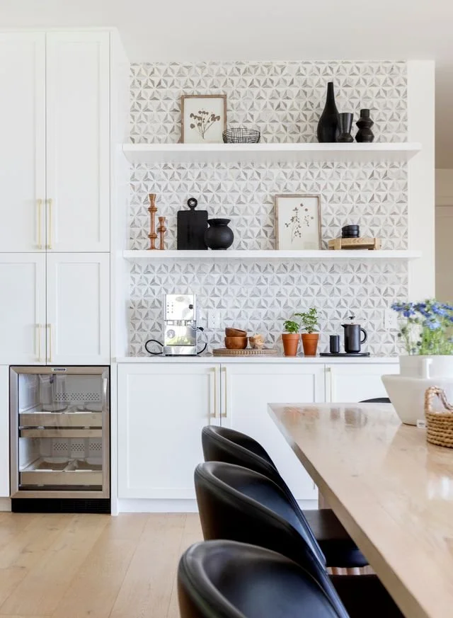 Modern kitchen with white cabinets, open shelves, decorative vases, framed botanical art, potted plants, and a dining table with black chairs.