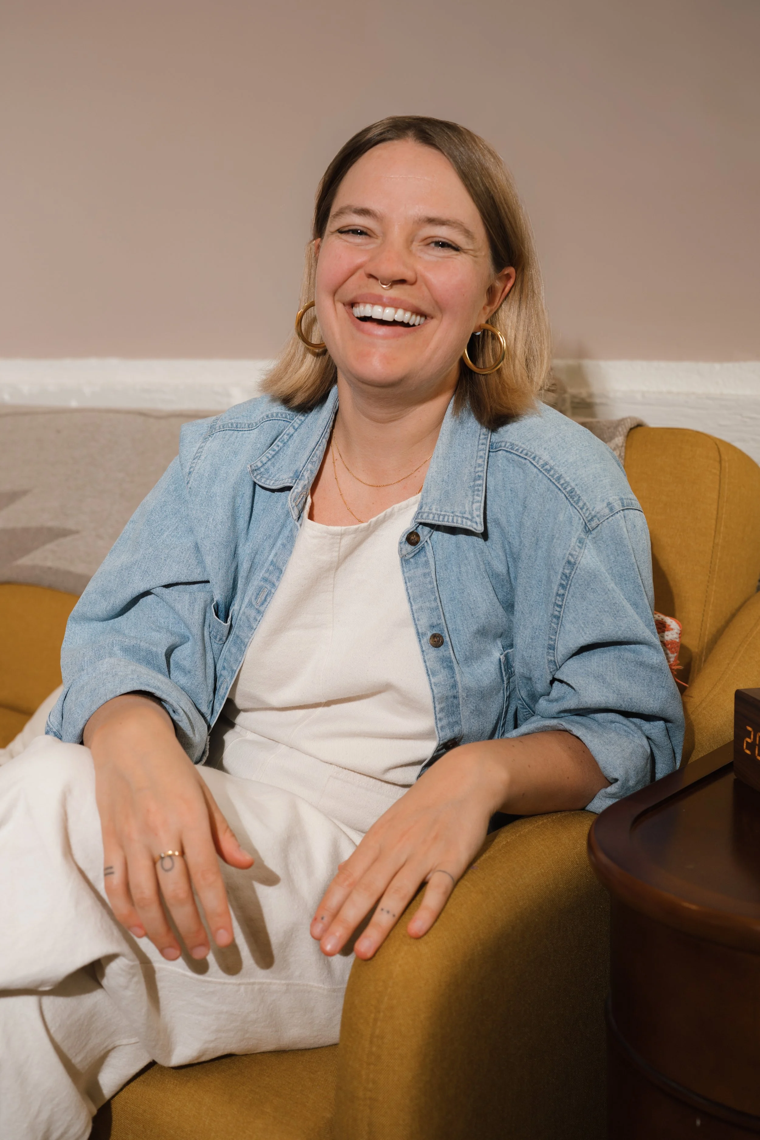 A woman sitting comfortably on a yellow armchair, smiling warmly, wearing a denim jacket over a white top, with jewelry and earrings, in a cozy indoor setting.