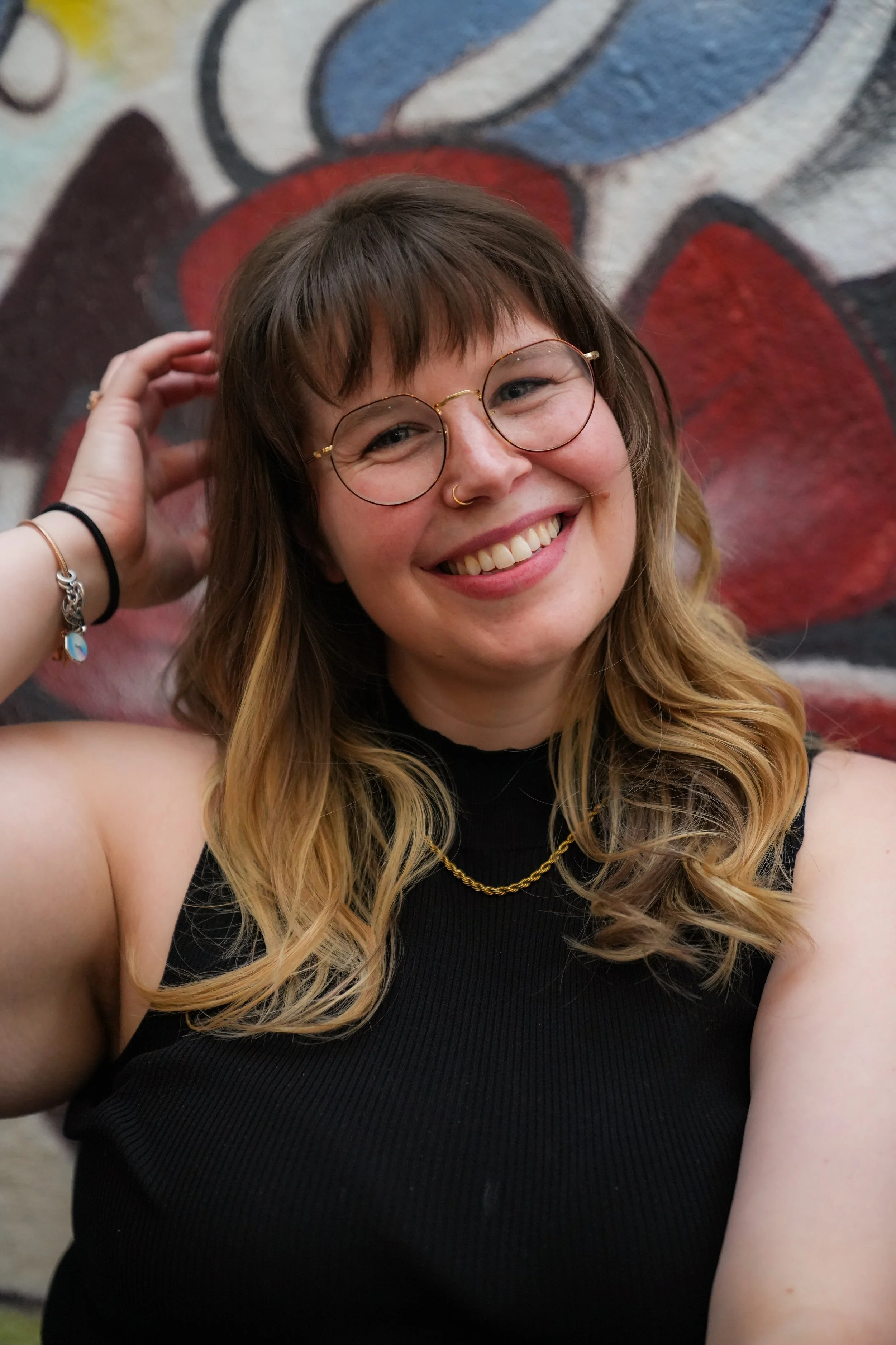 A young woman with glasses, a nose ring, and wavy blonde hair, smiling and looking at the camera, standing in front of a colorful graffiti wall.