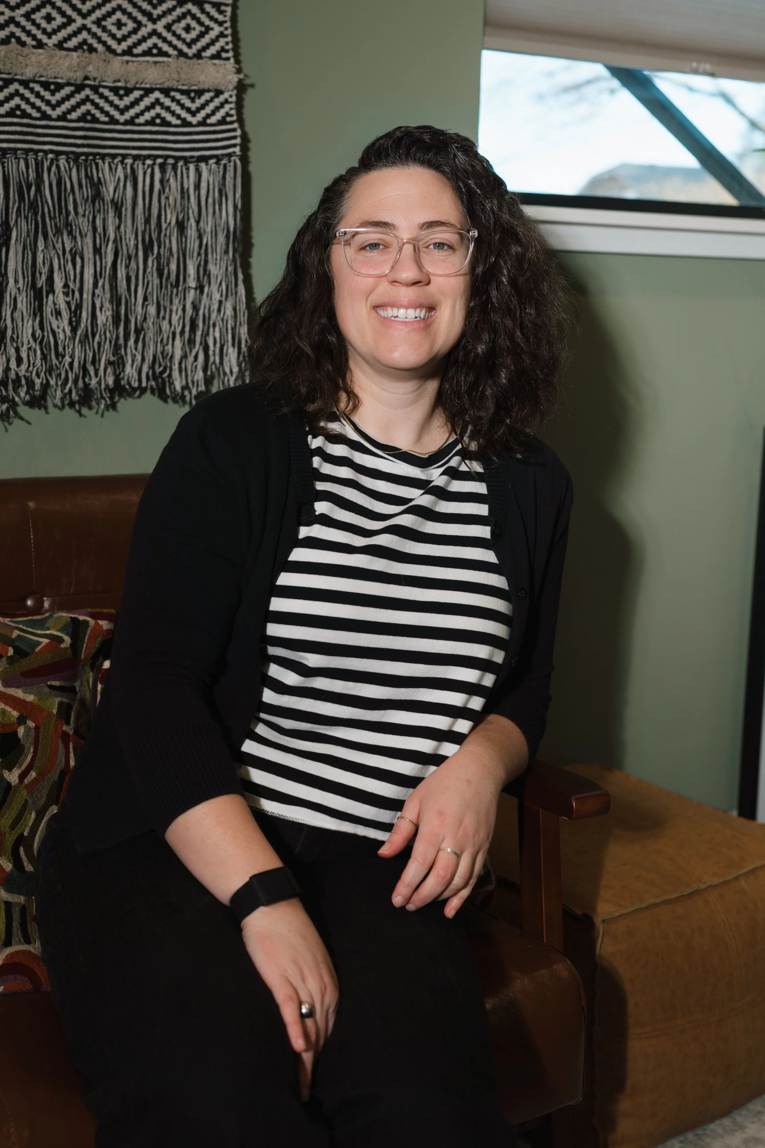 A woman with dark curly hair, glasses, and a black and white striped shirt sits on a leather chair in a room with green walls and a window in the background.