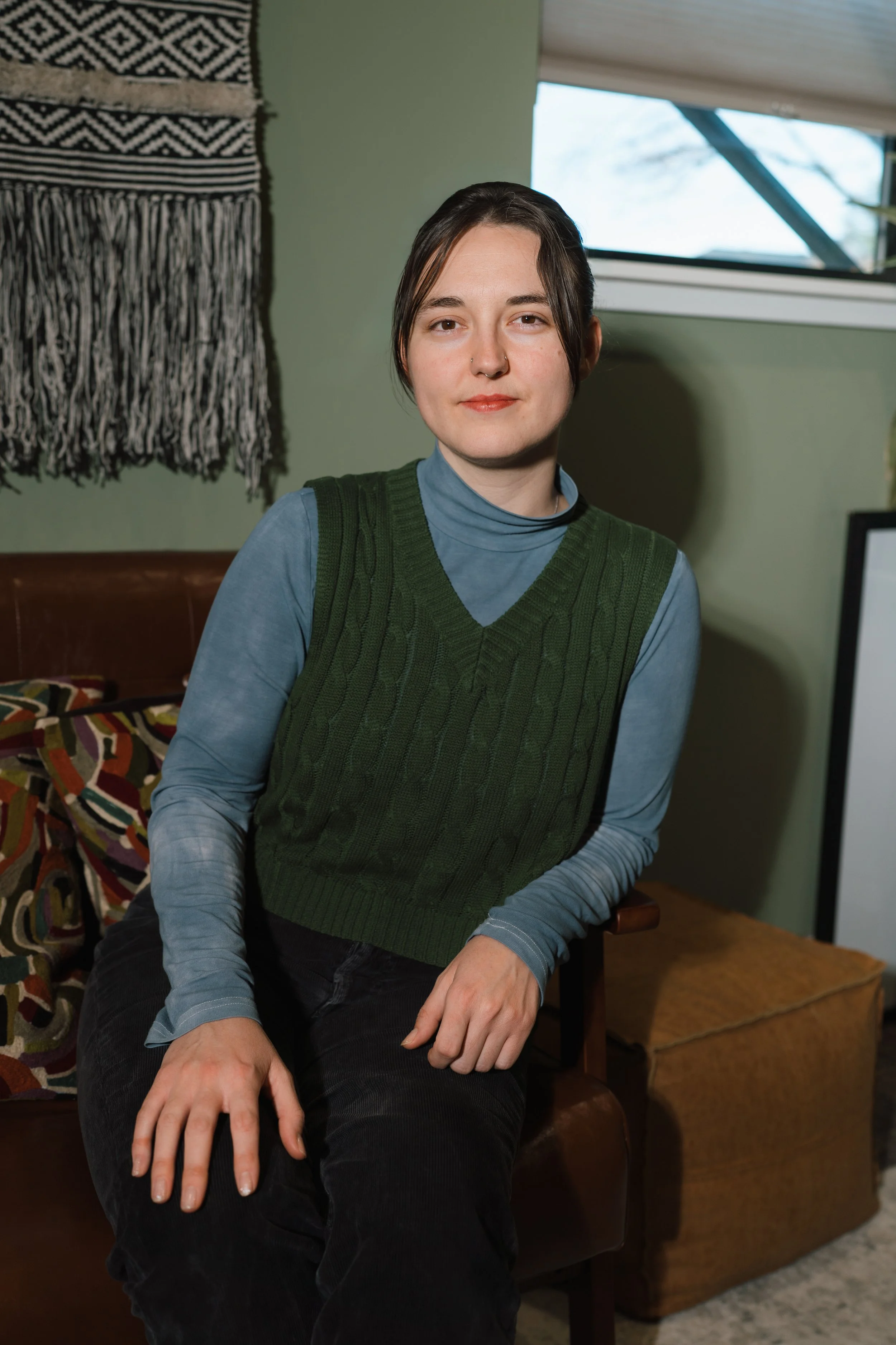 A young woman with short dark hair, wearing a green cable-knit vest over a blue long-sleeve turtleneck, sitting on a brown chair in a room with green walls, a patterned blanket hanging on the wall, and a window with a light-colored shade partially open behind her.
