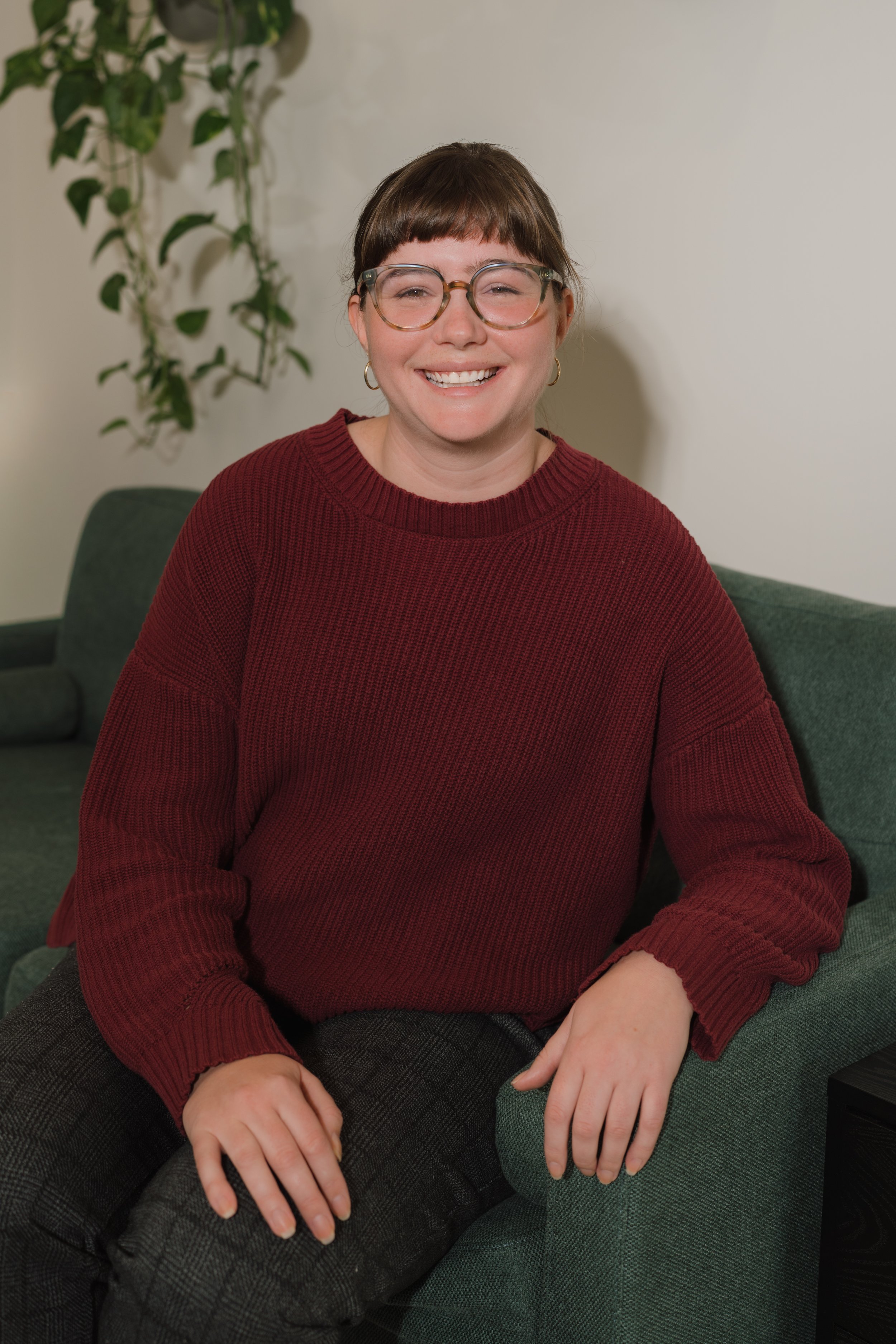 A woman with short brown hair, glasses, and earrings, smiling, wearing a red sweater and sitting on a green sofa in a room with a plant behind her.