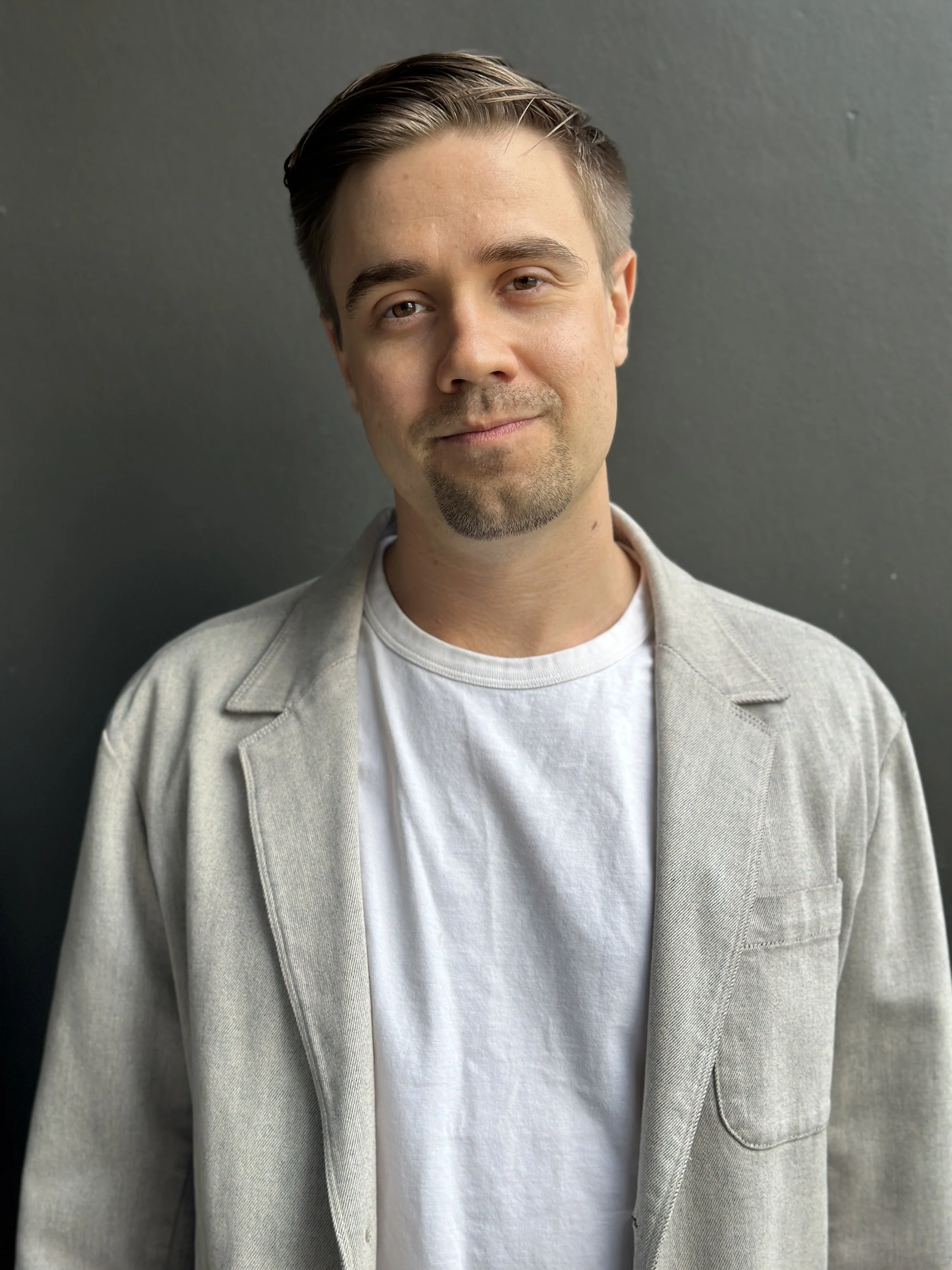 A young man with light skin, short brown hair, and a light beard and mustache, wearing a light gray blazer over a white t-shirt, standing against a dark gray wall, smiling slightly.