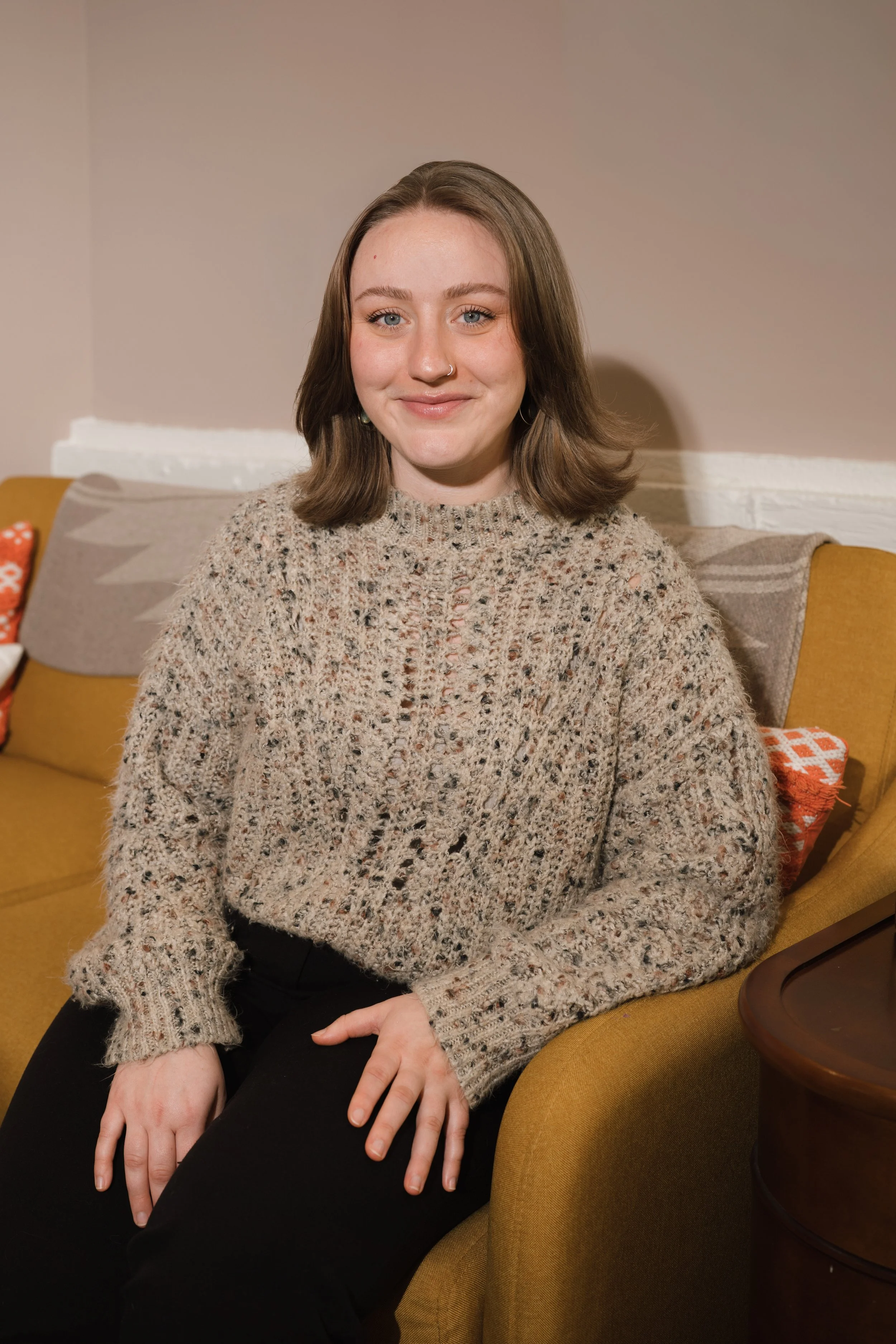 A woman with shoulder-length brown hair and blue eyes sitting on a mustard yellow couch, wearing a chunky beige knit sweater.