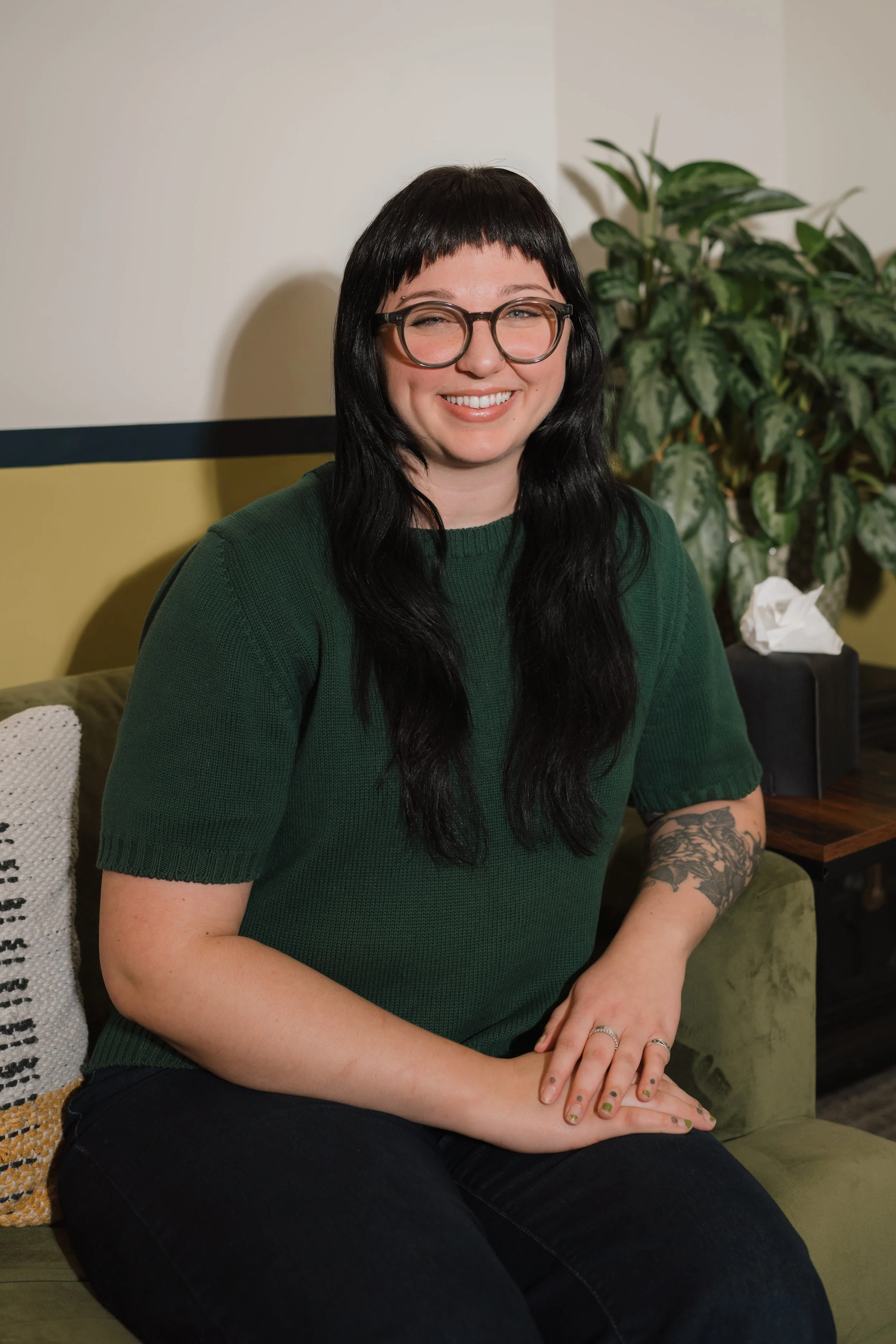 A woman with long black hair, glasses, and tattoos, wearing a green sweater, sitting on a green sofa in a room with a plant and a tissue box behind her.