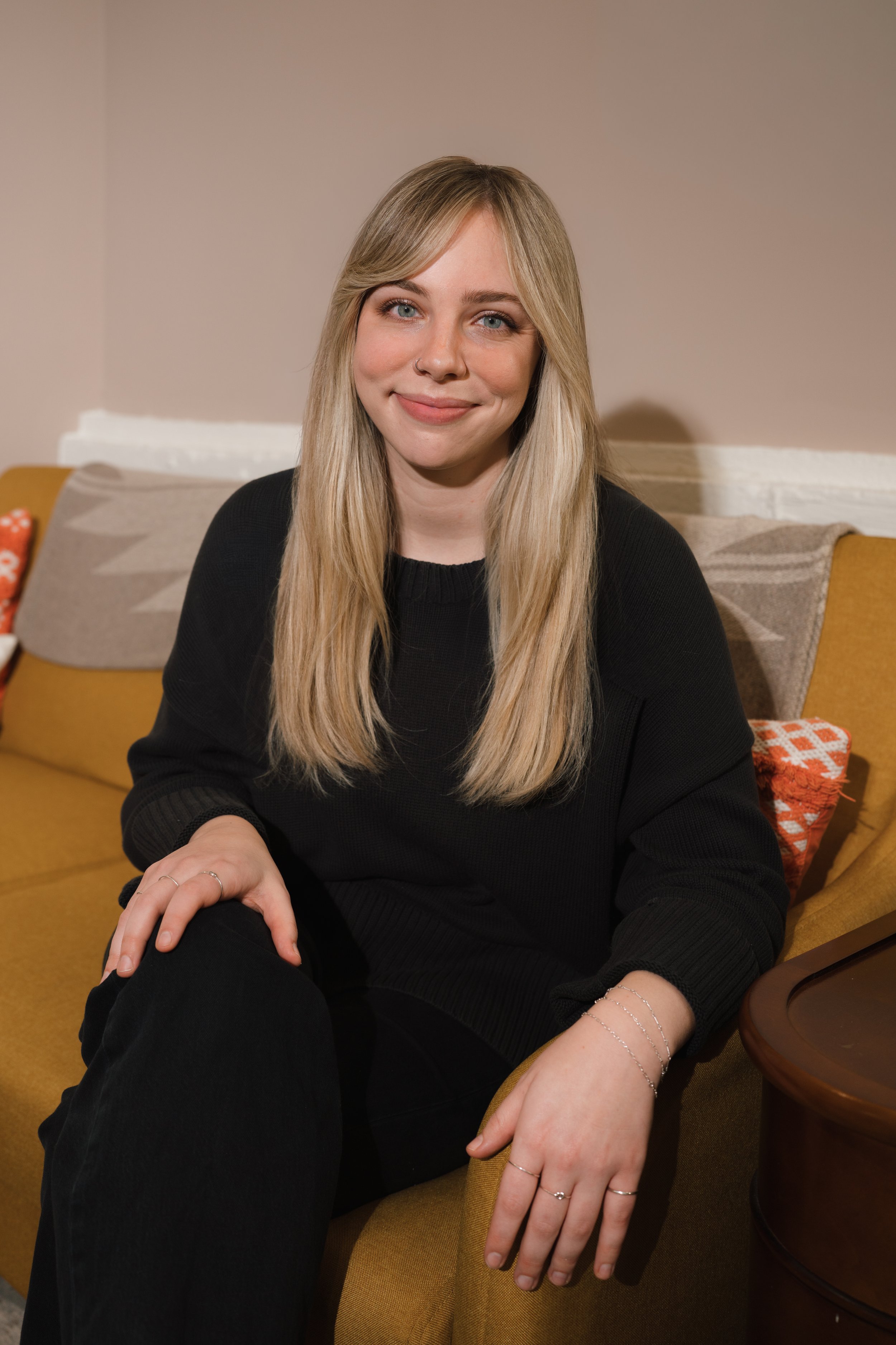 A young woman with long blonde hair and blue eyes, wearing a black sweater, sitting on a yellow couch in a cozy room.