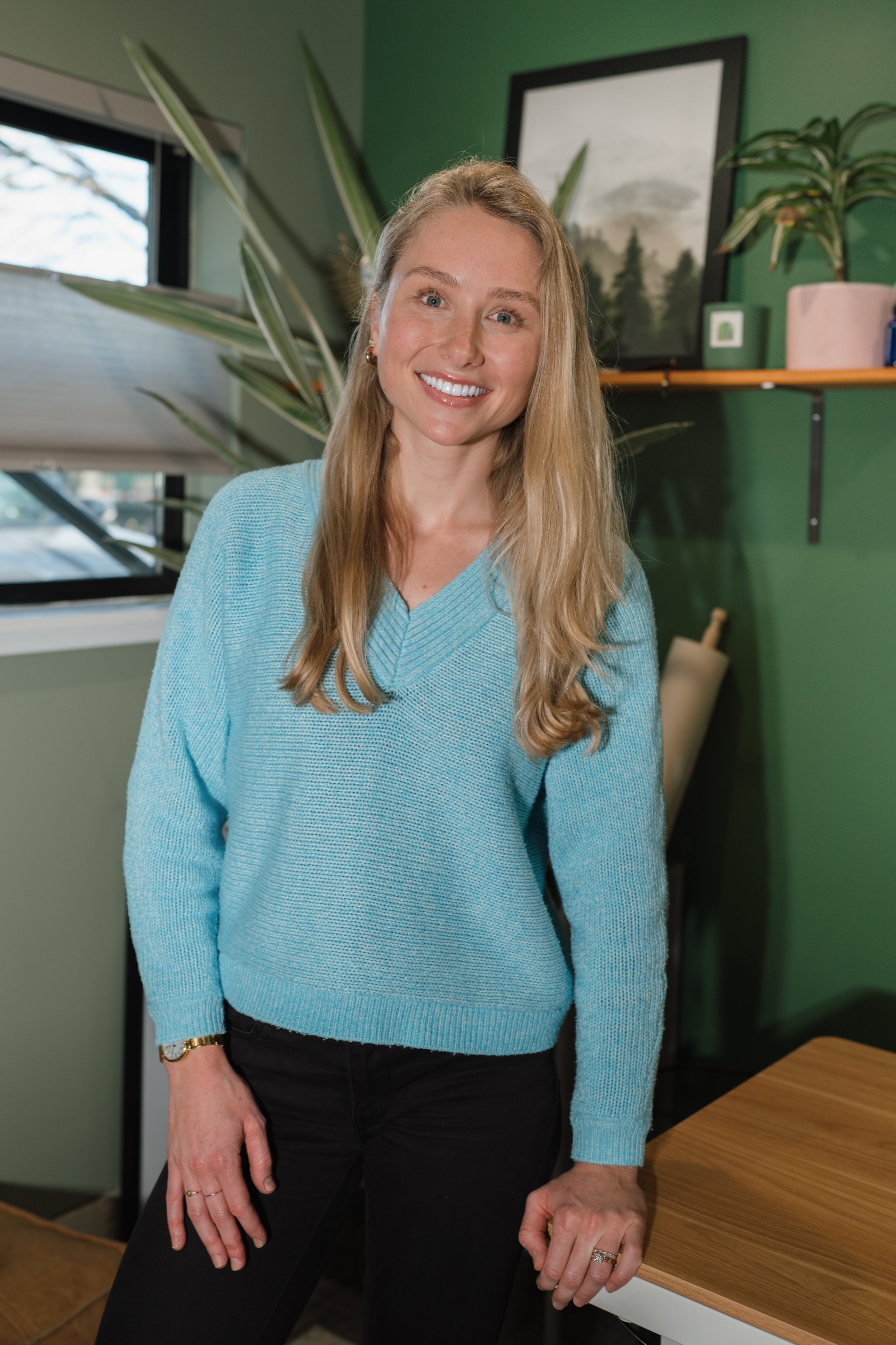 A woman with long blonde hair, smiling, wearing a light blue sweater and black pants, standing indoors next to a table with a wooden top, with a green wall, bookshelf, and window in the background.