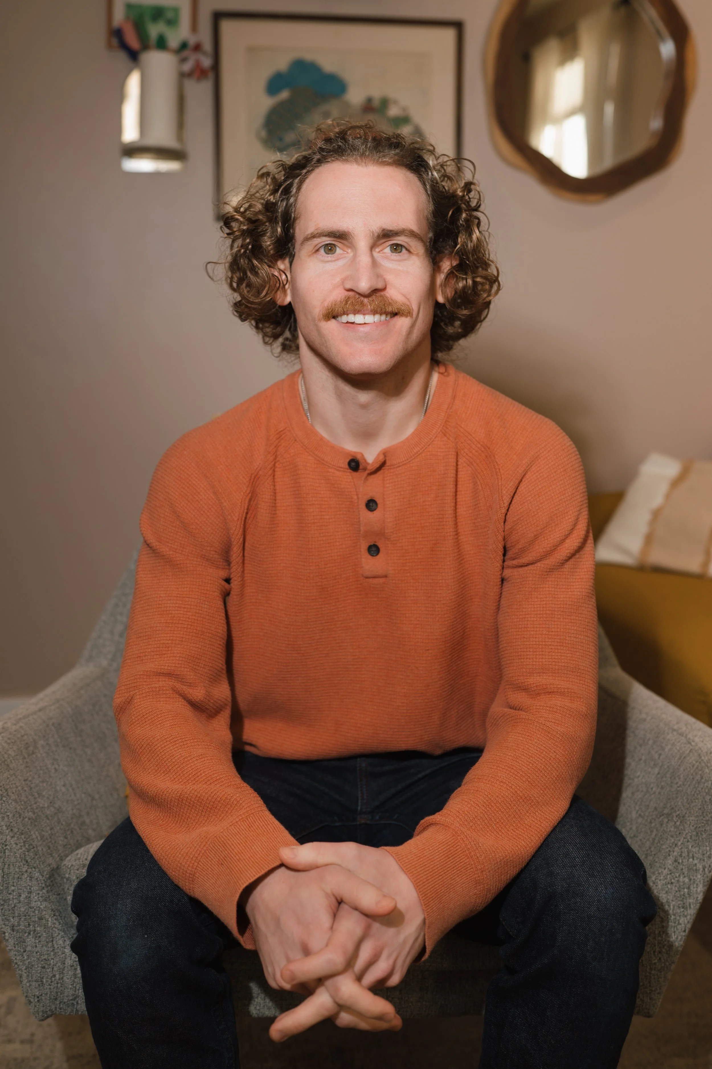 A man with curly hair and a mustache, wearing an orange long-sleeved shirt, sitting on a beige armchair in a cozy room.