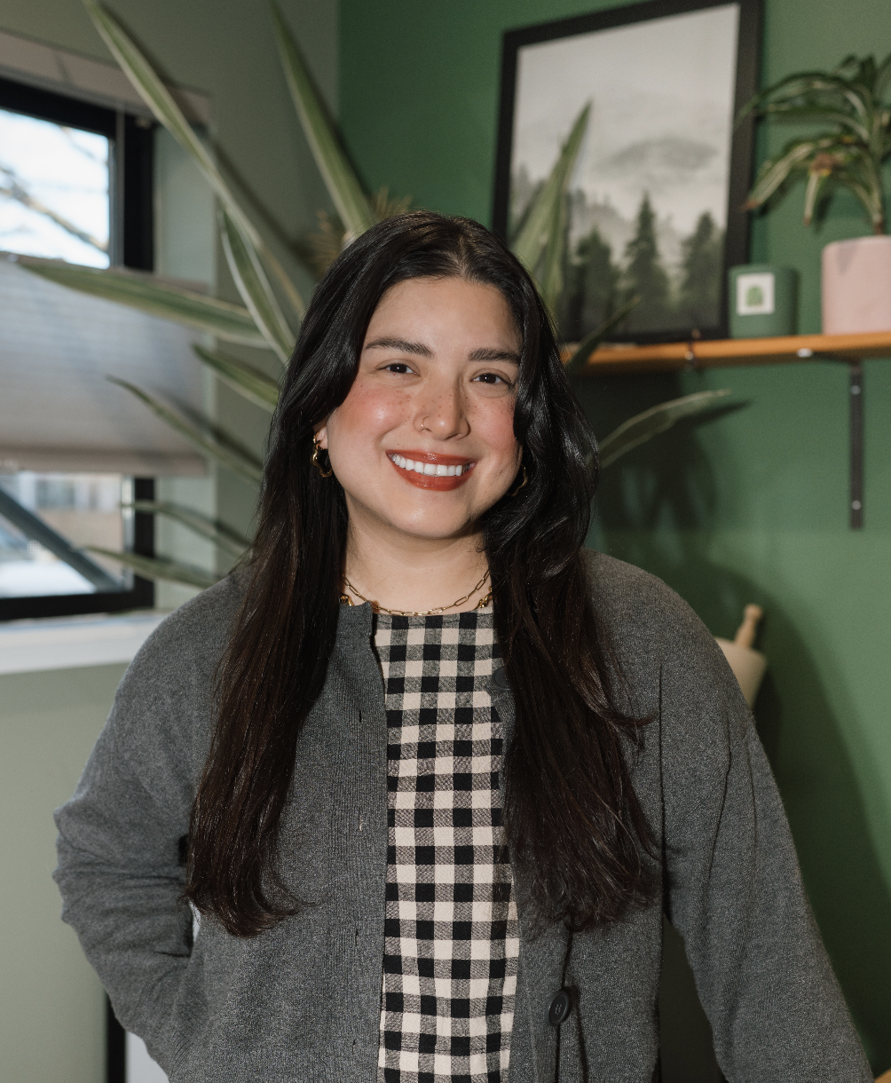 A woman with long dark hair and a smile, wearing a checkered top and gray blazer, stands in a green-walled room with plants and a window.