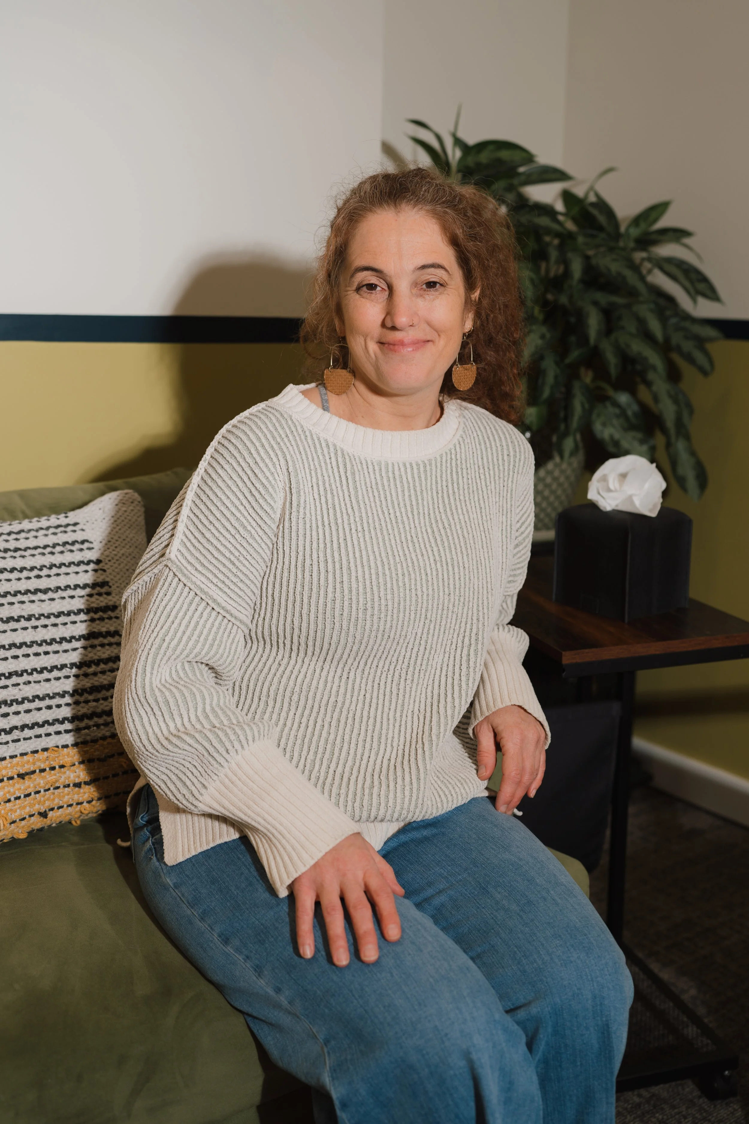 A woman sitting on a green sofa, smiling at the camera, wearing a cream sweater and blue jeans, with a decorative pillow and a tissue box on a side table behind her.