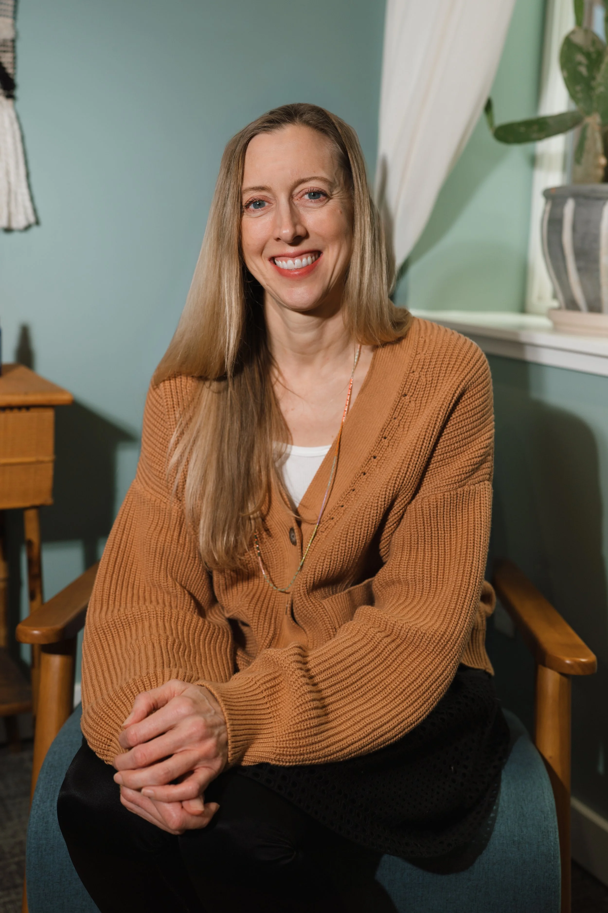 A woman with long blonde hair, smiling, wearing a brown cardigan over a white top, sitting on a blue chair in a room with green walls, a window with white curtains, and a potted plant in the background.
