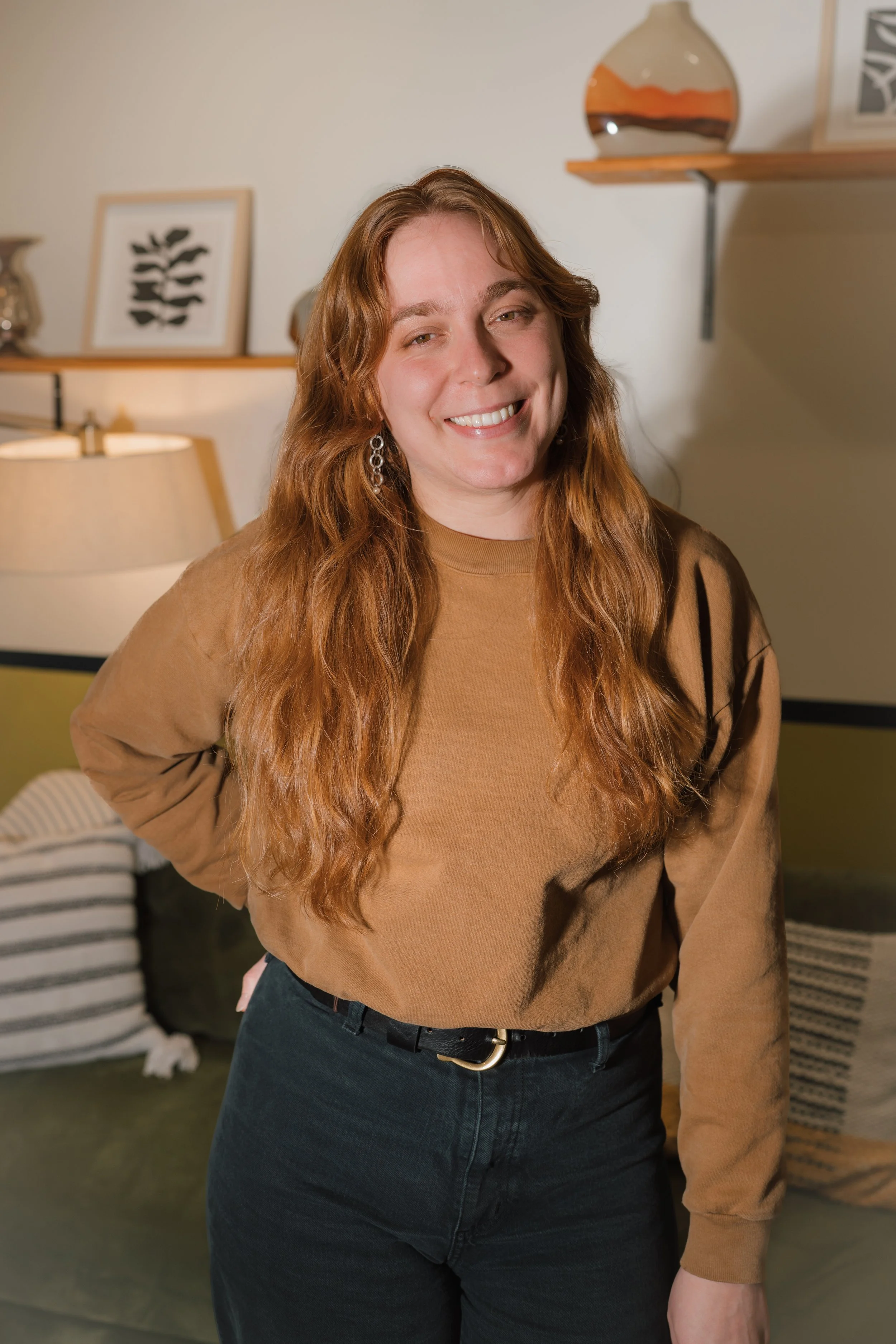 A woman with long, wavy red hair smiling in a cozy indoor setting.