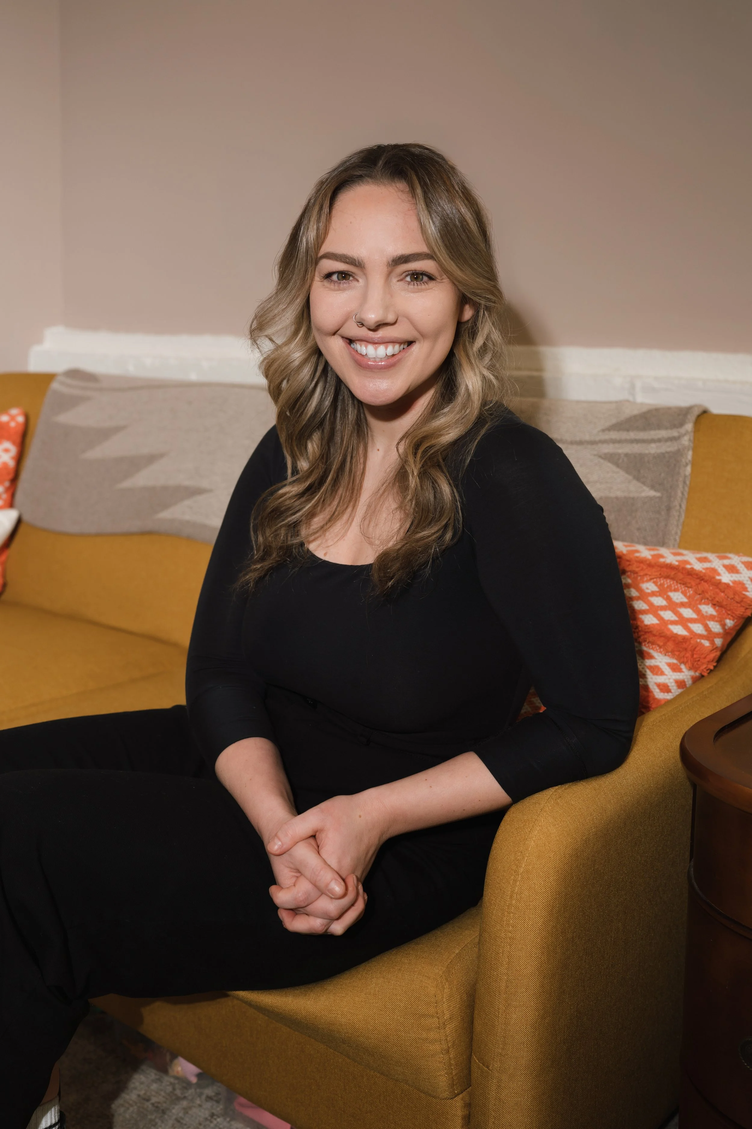 A young woman with wavy blonde hair, smiling, sitting on a mustard-colored armchair in a living room.