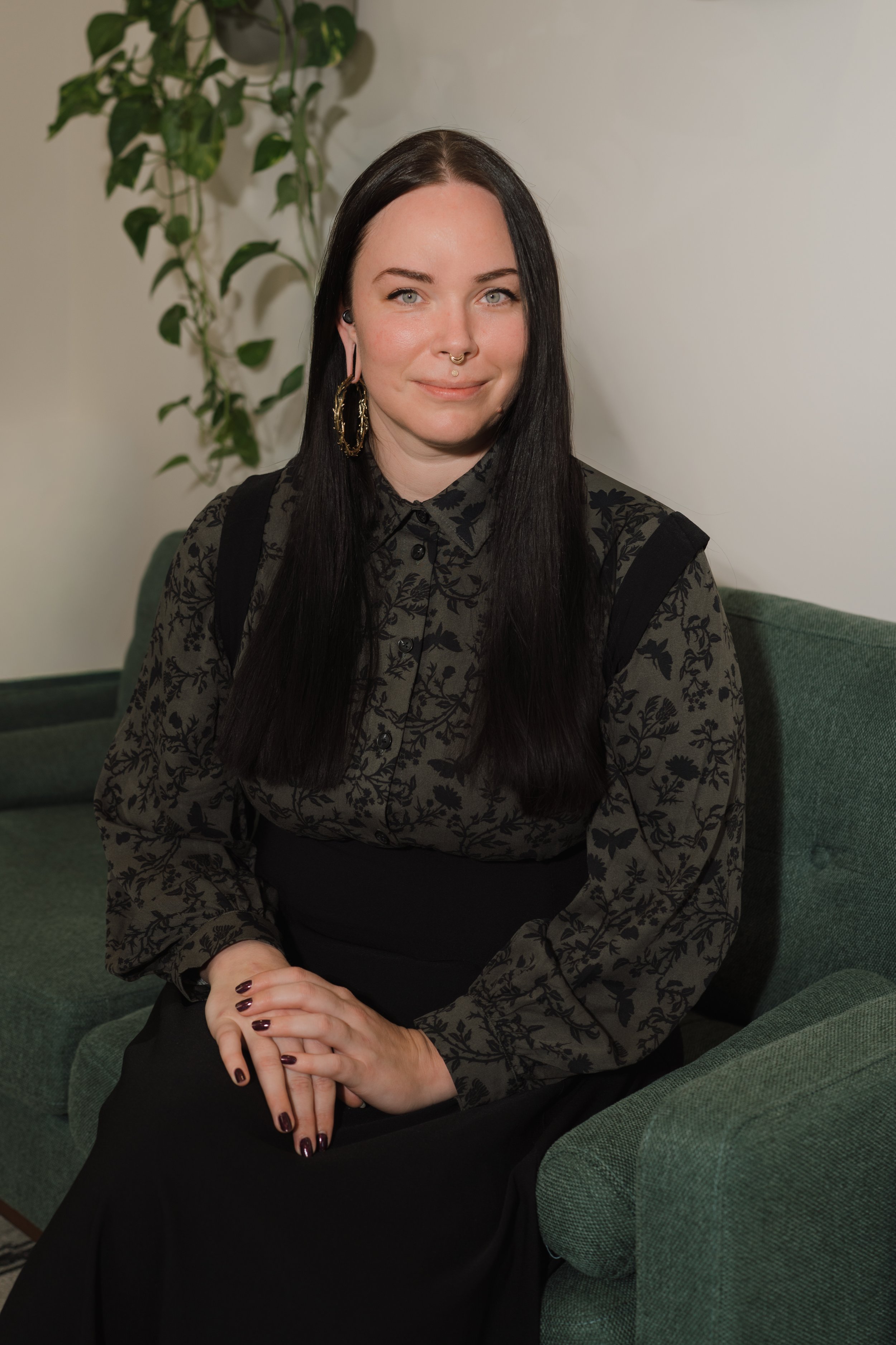 A young woman with long black hair, wearing a black and gray floral blouse, sitting on a green couch with her hands clasped, in front of a white wall with a leafy green plant.