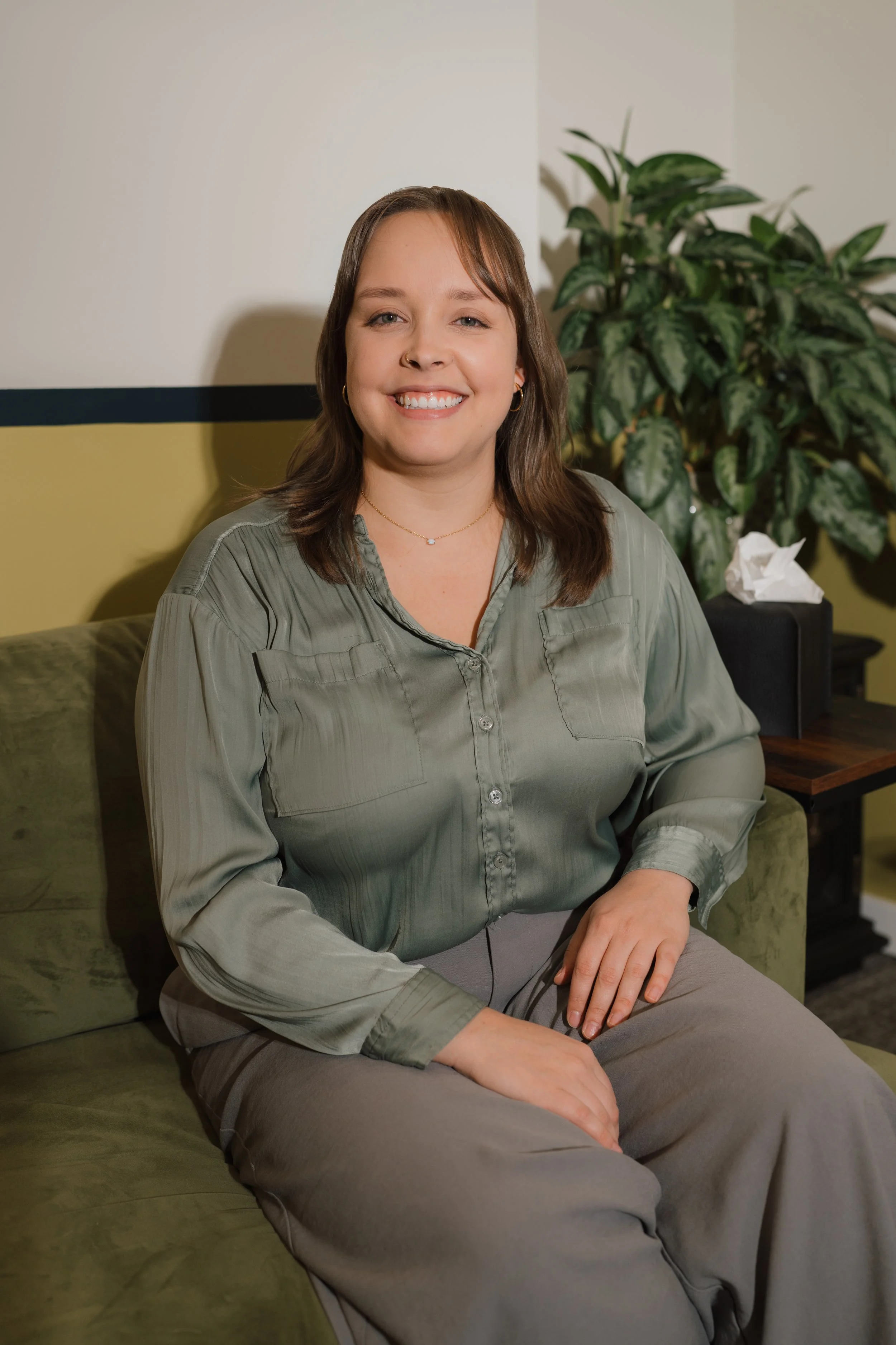 A young woman with shoulder-length brown hair, wearing a light green button-up shirt and gray pants, sitting on a green sofa in a room with a large leafy plant and a tissue box on a small table behind her, smiling at the camera.