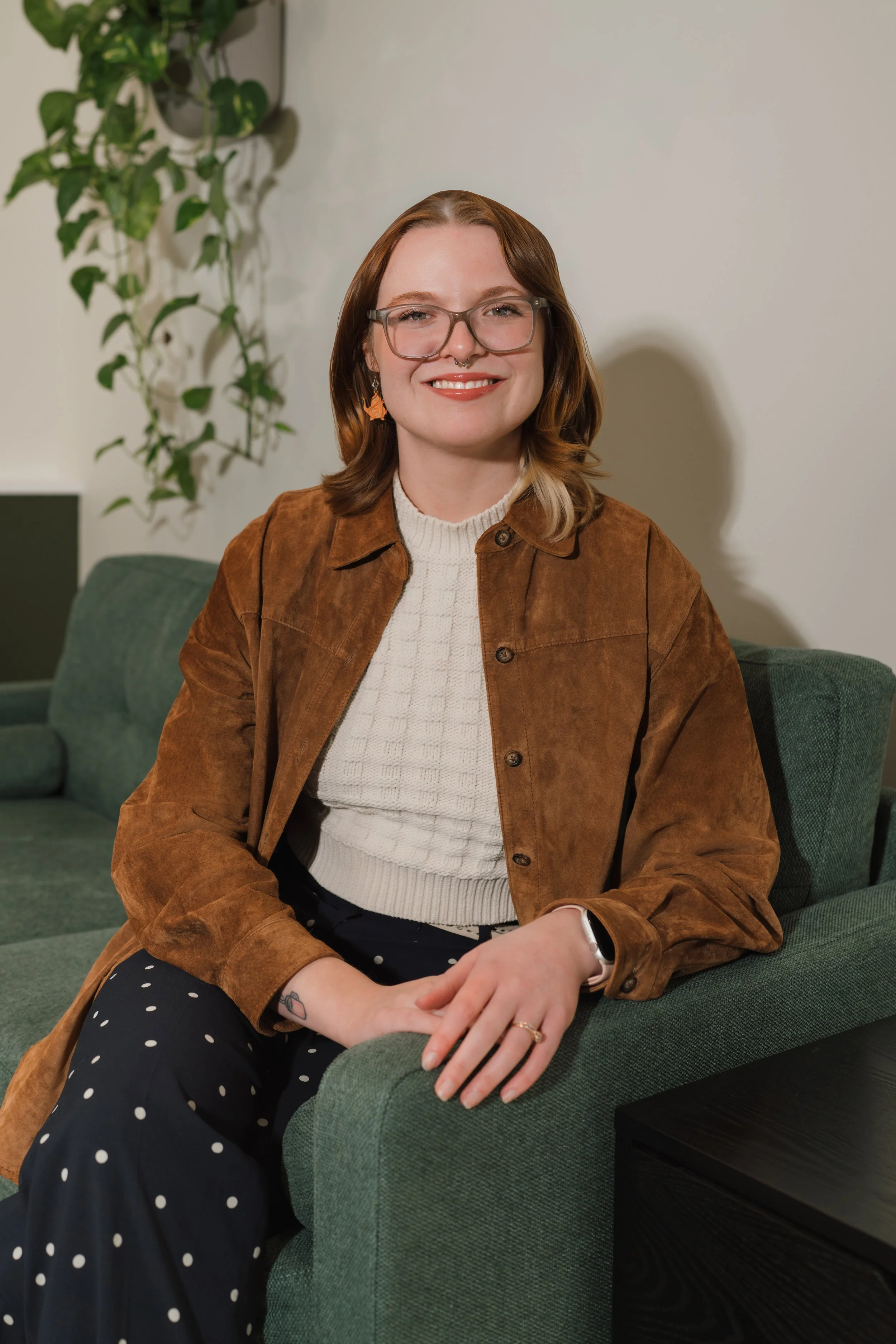 A woman with shoulder-length brown hair, glasses, and a septum piercing smiling while sitting on a green couch in an indoor setting with a plant in the background.
